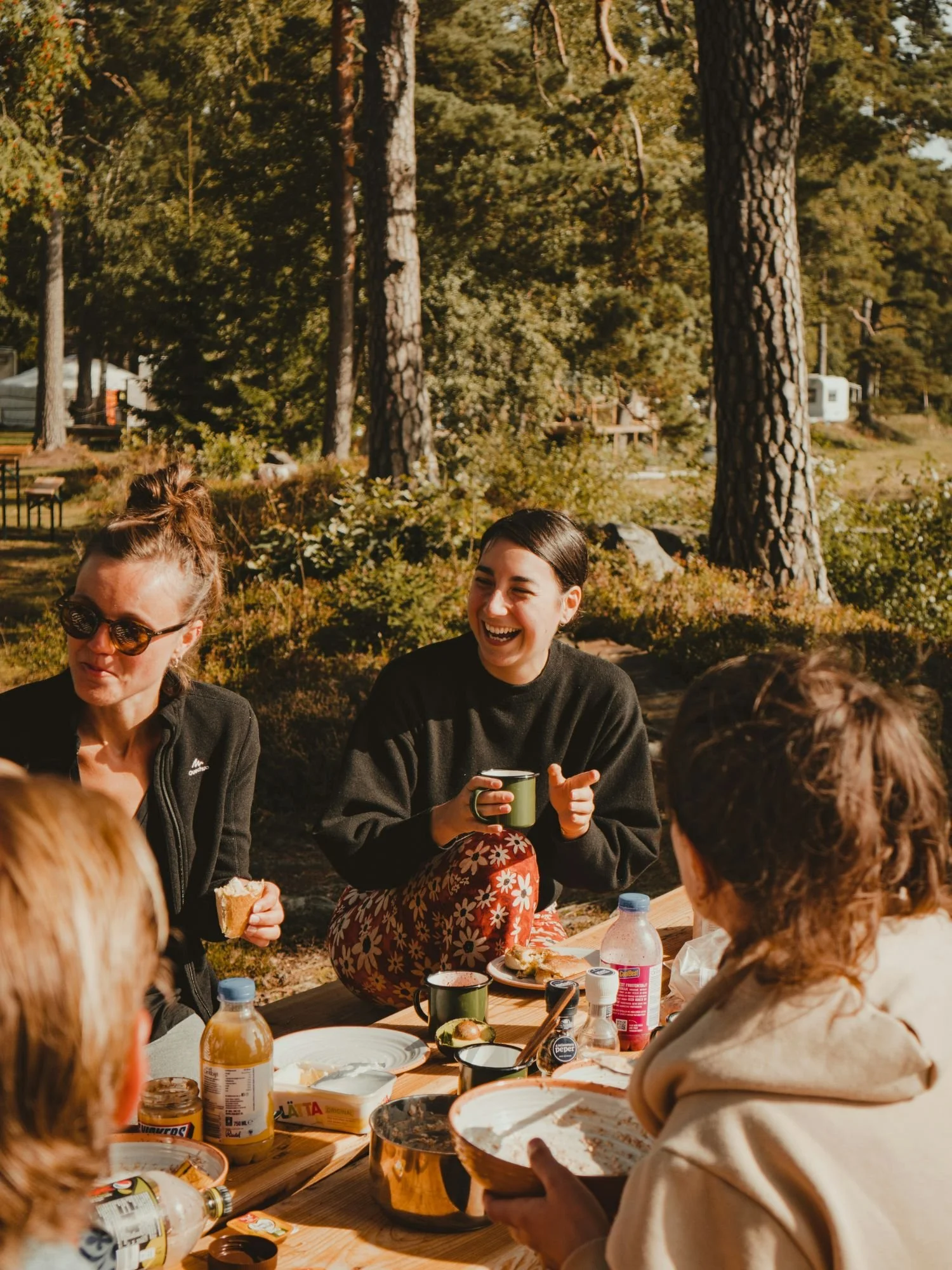 A group of people are gathered around a wooden picnic table enjoying an outdoor breakfast in a sunlit, wooded campground.