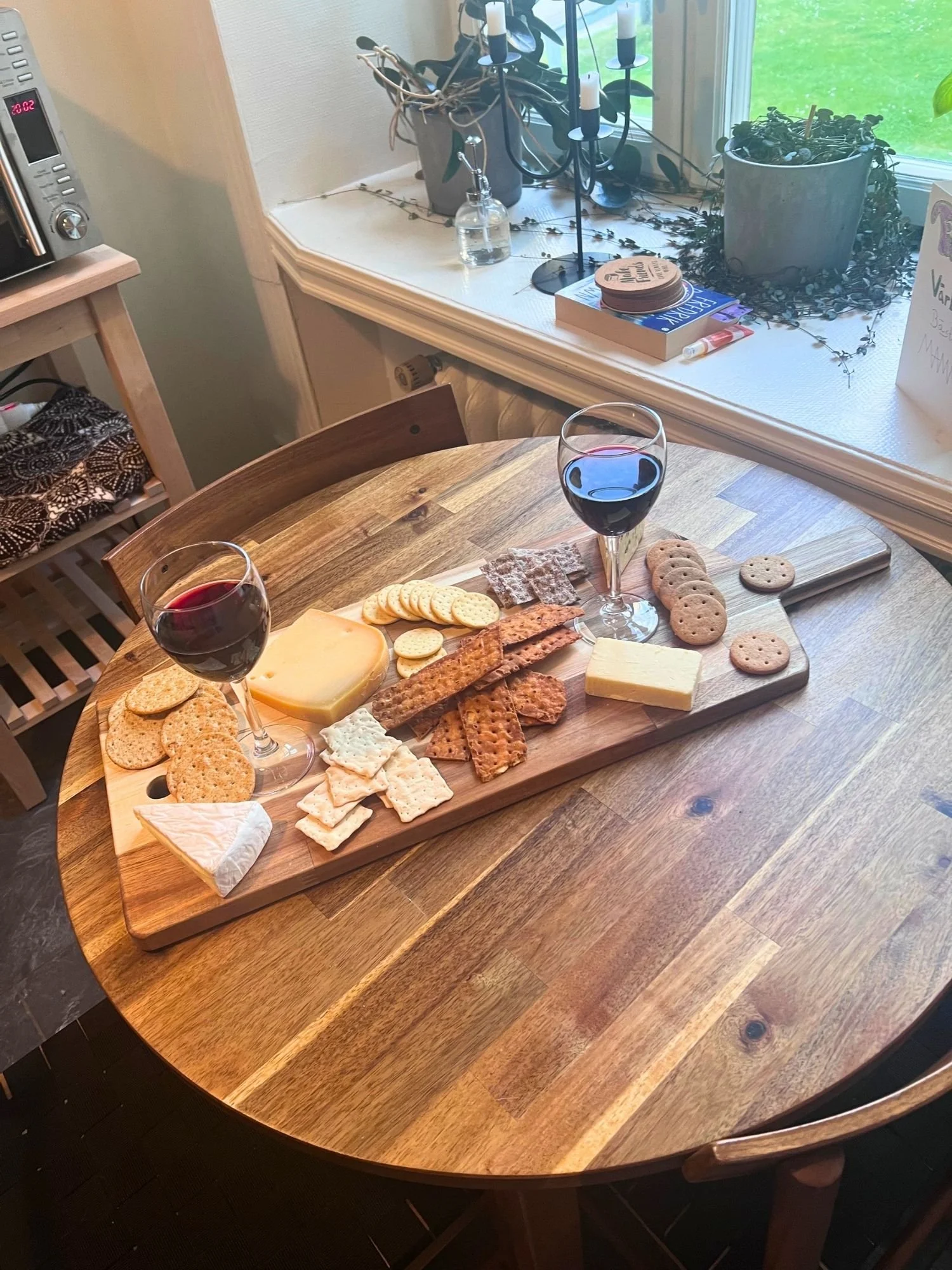 A wooden charcuterie board on a round wooden table filled with an assortment of crackers and cheeses, accompanied by two glasses of red wine.