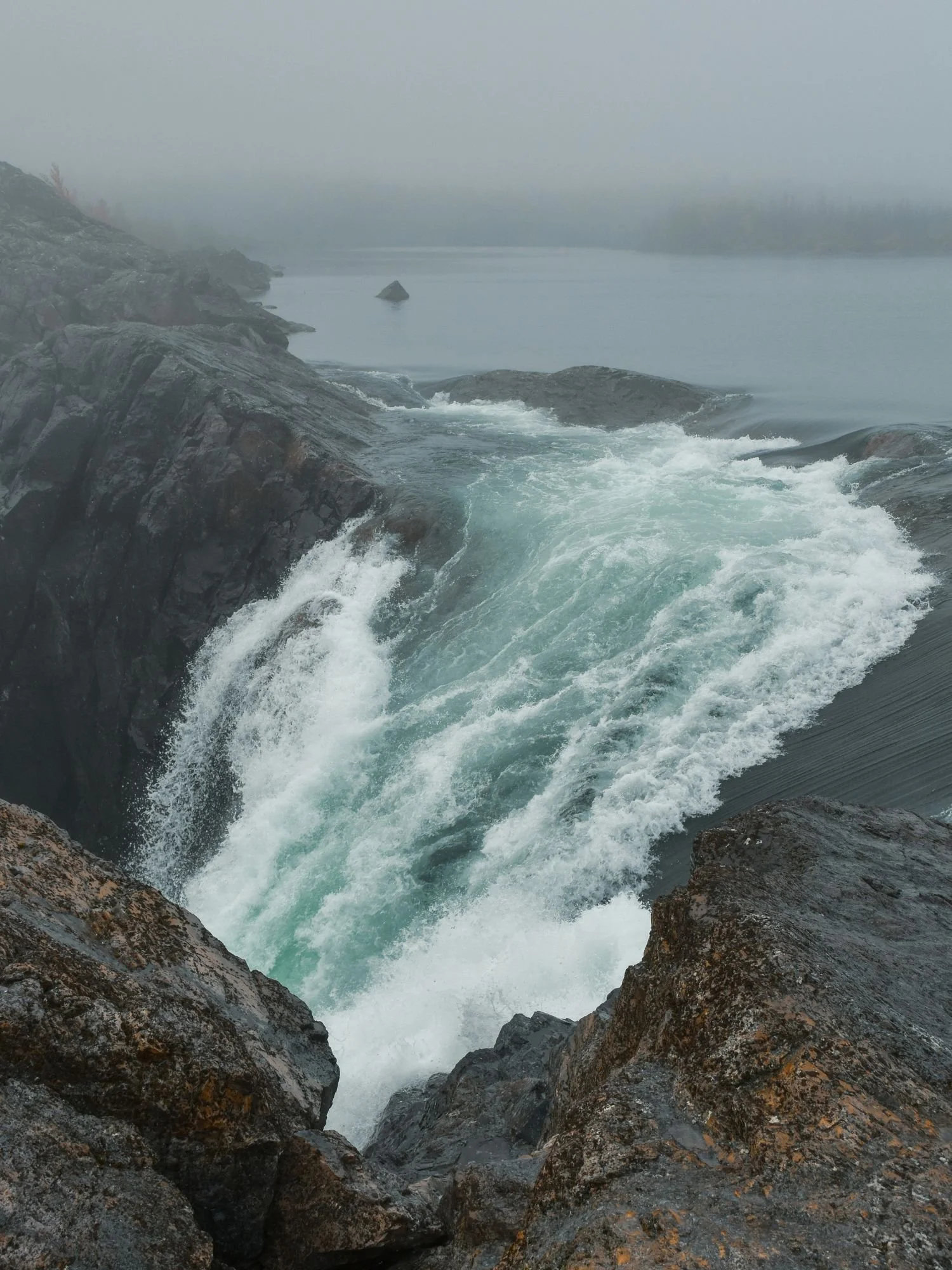 A powerful, churning turquoise waterfall flows between dark, rocky cliffs under a thick blanket of fog in Stora Sjöfallet National Park.