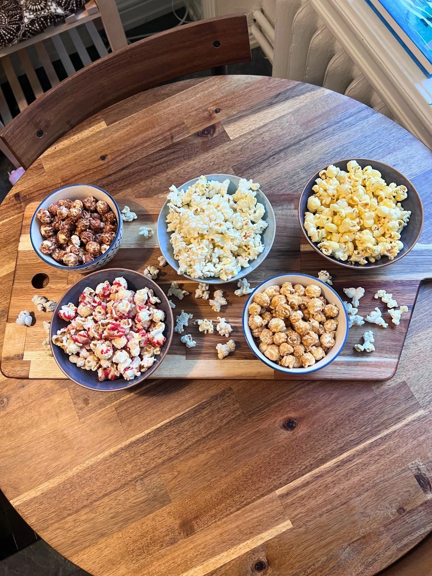 High-angle, top-down view of five ceramic bowls filled with different flavors of gourmet popcorn arranged on a wooden board on a round wooden table.