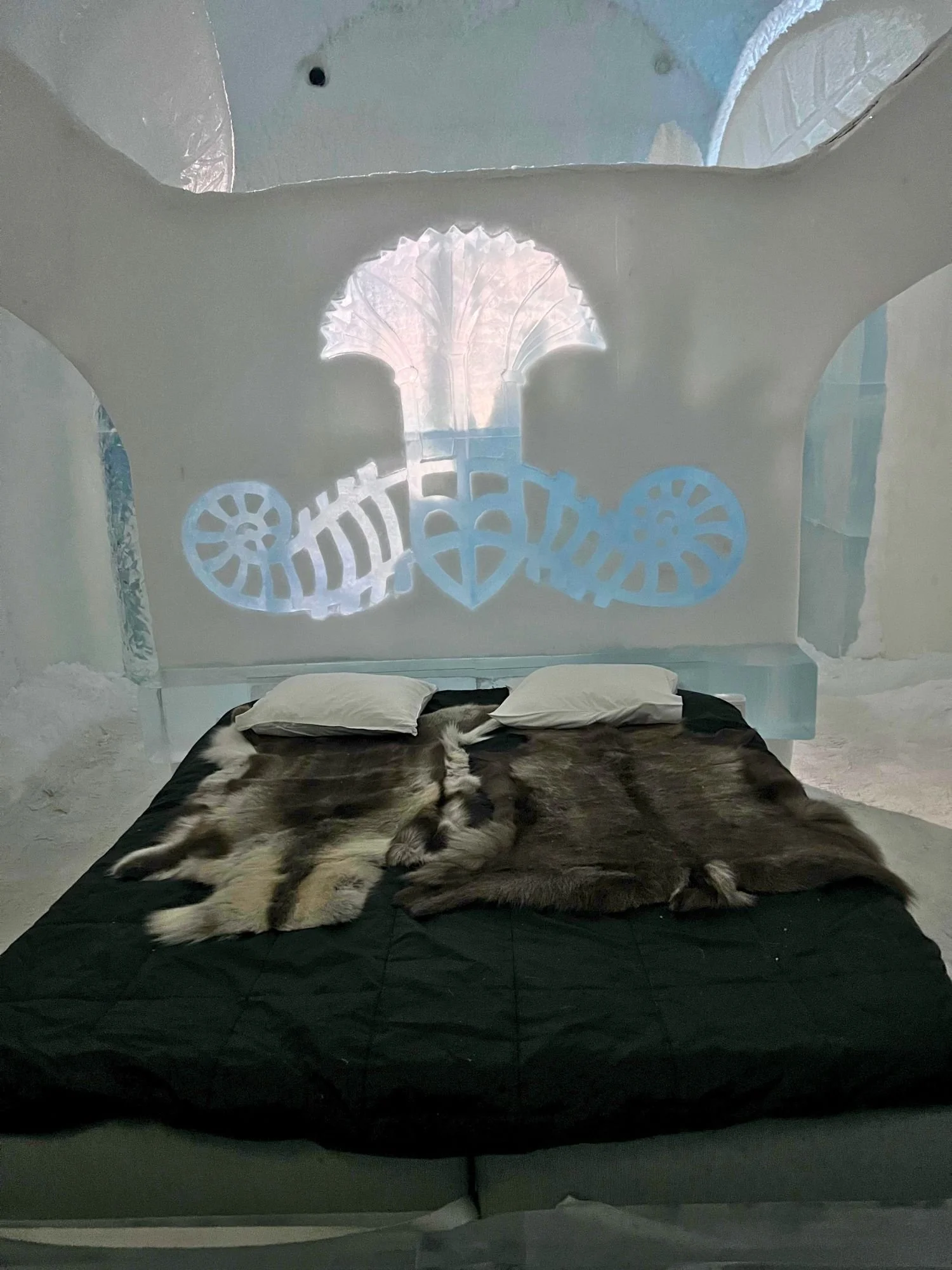 Ice room at Sweden’s Icehotel featuring a carved ice headboard with illuminated artwork, a bed covered in reindeer hides, and frosty white surroundings.