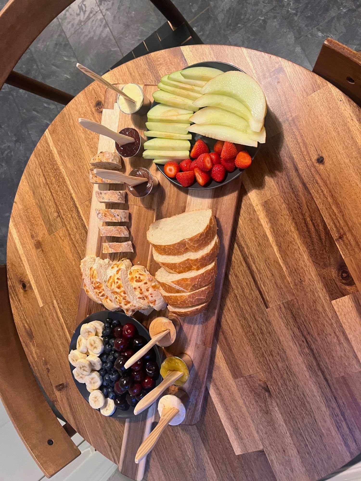 High-angle shot of a long wooden serving board on a round wooden table, arranged with sliced bread, plates of fresh fruit including bananas, berries, melon, and strawberries, and small cups of spreads with wooden spoons.