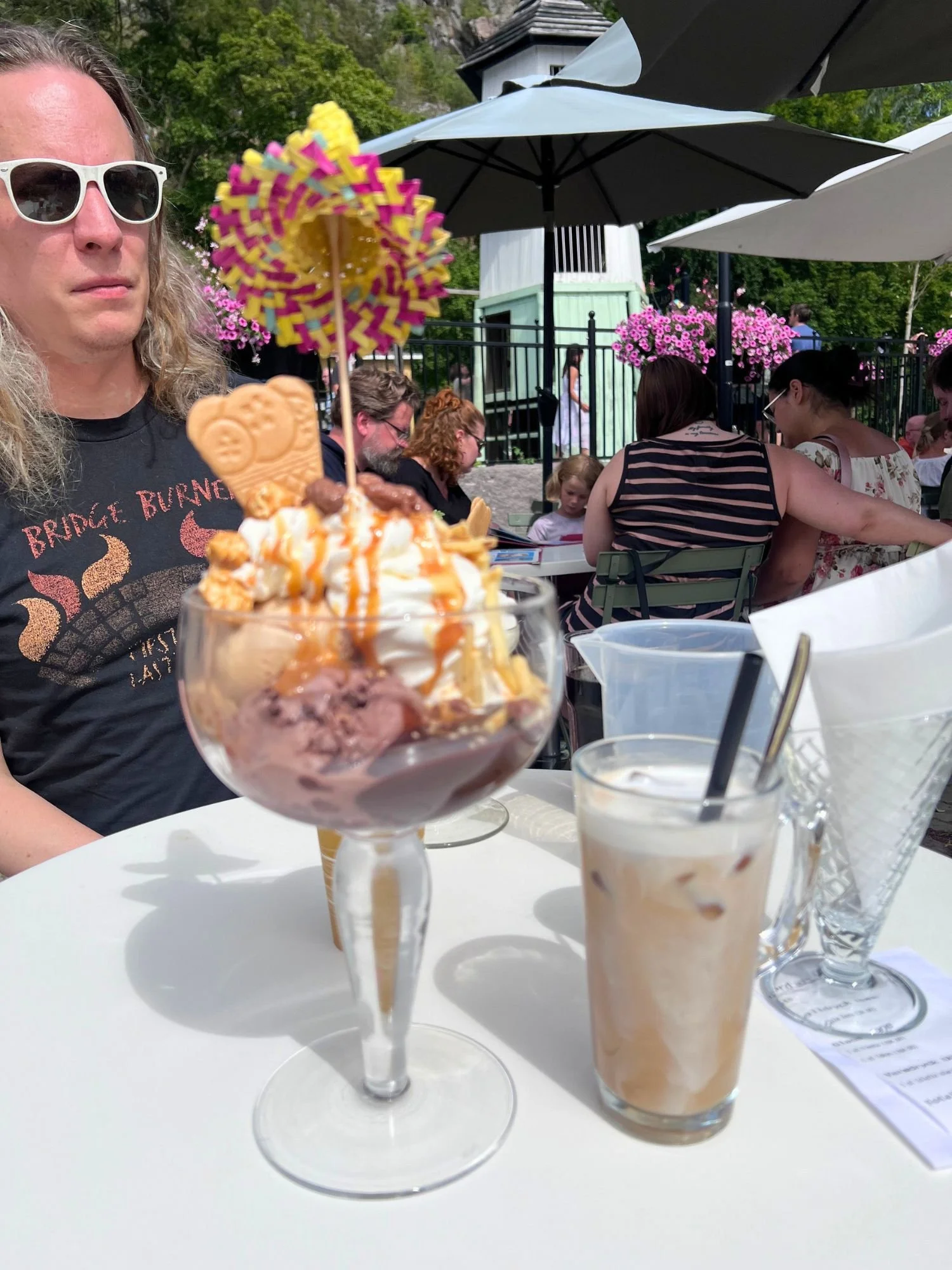An elaborate chocolate and caramel ice cream sundae topped with a miniature woven sombrero alongside an iced latte on an outdoor restaurant table.