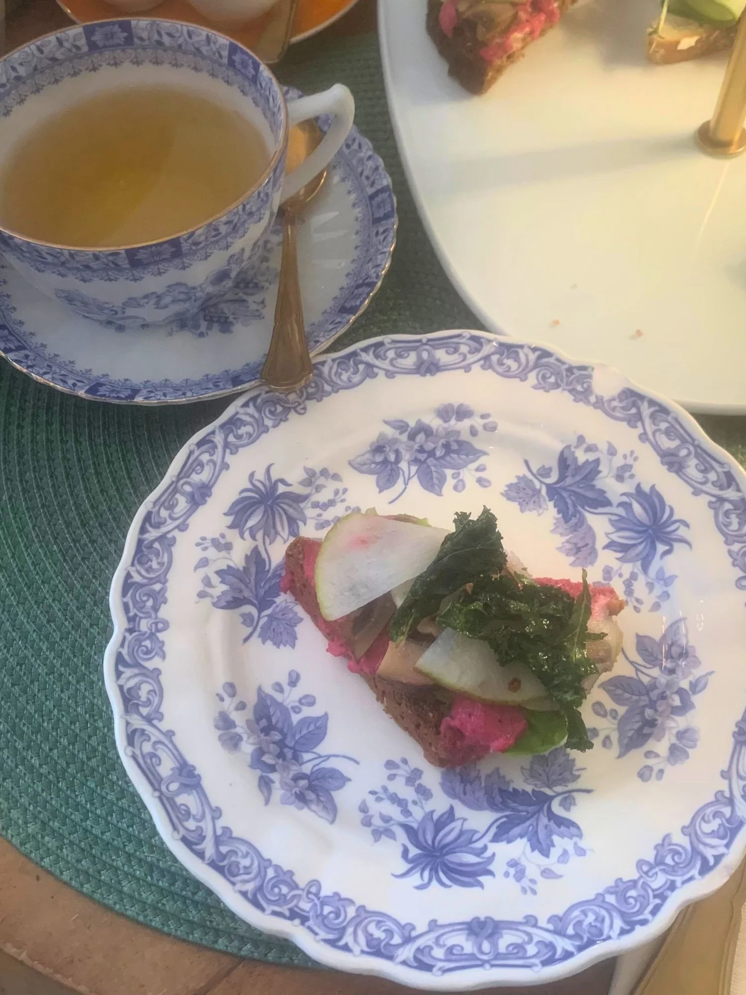 An open-faced sandwich and tea on a table at Alice och Kattens Tesalong in Lund. A piece of dark bread topped with a bright pink spread, sliced radishes, and dark leafy greens on a white plate with an intricate blue floral pattern.