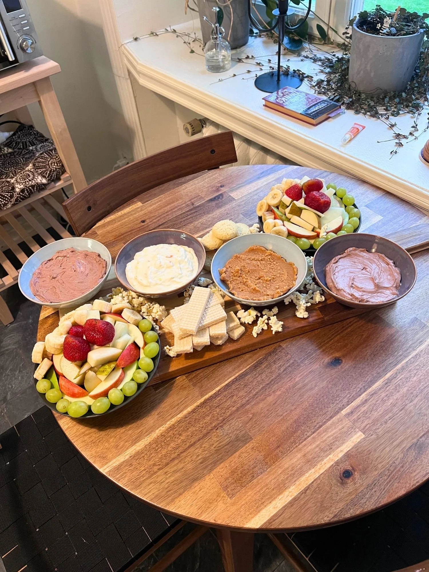 A wooden table set with a snack platter featuring four bowls of sweet dessert dips, two plates of fresh fruit, vanilla wafer cookies, and scattered popcorn.