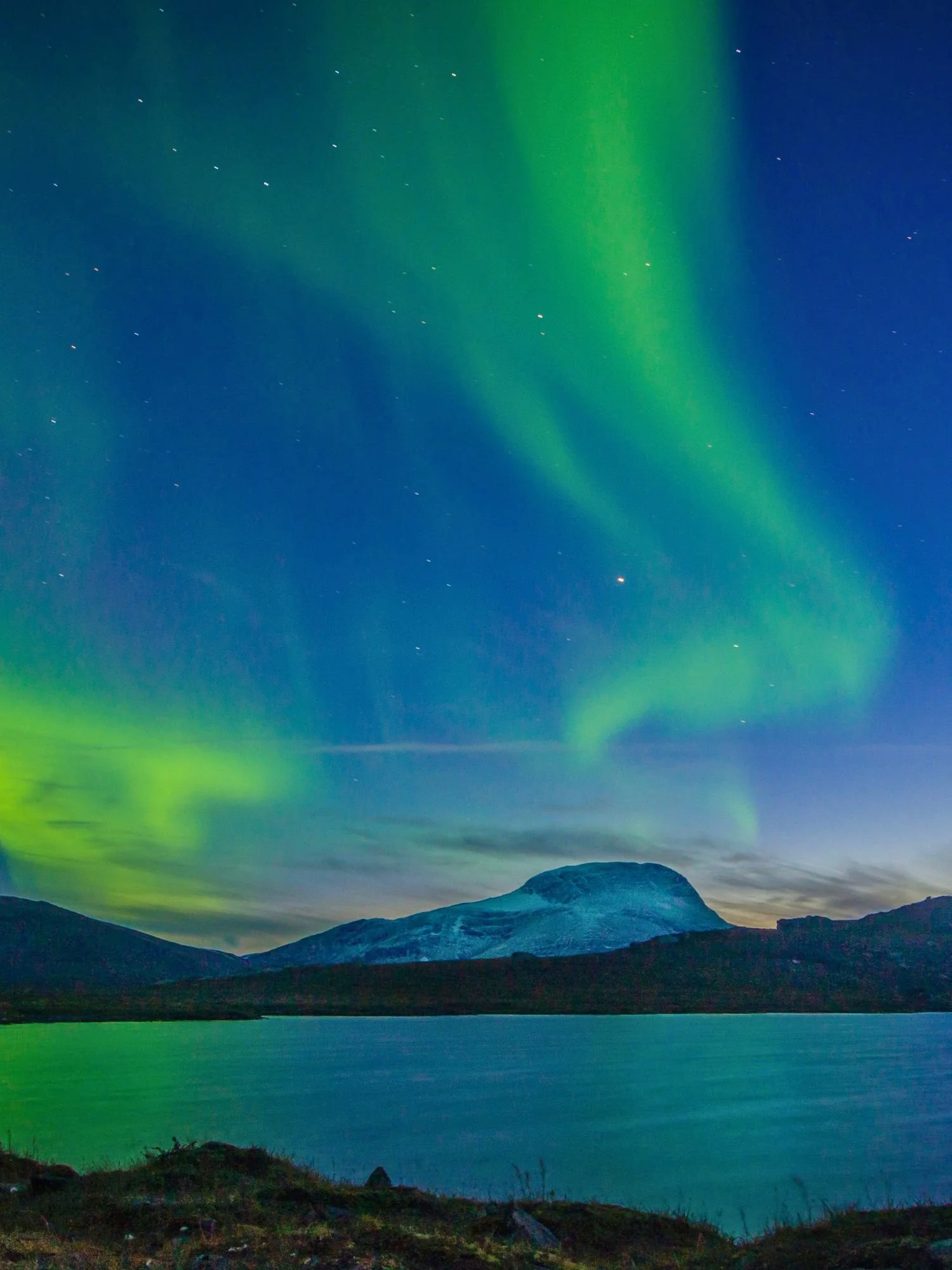 Vibrant green ribbons of the Aurora Borealis (Northern Lights) dancing across a deep blue, star-speckled night sky. A snow-dusted, dome-shaped mountain sits prominently behind a dark, low-lying ridge.