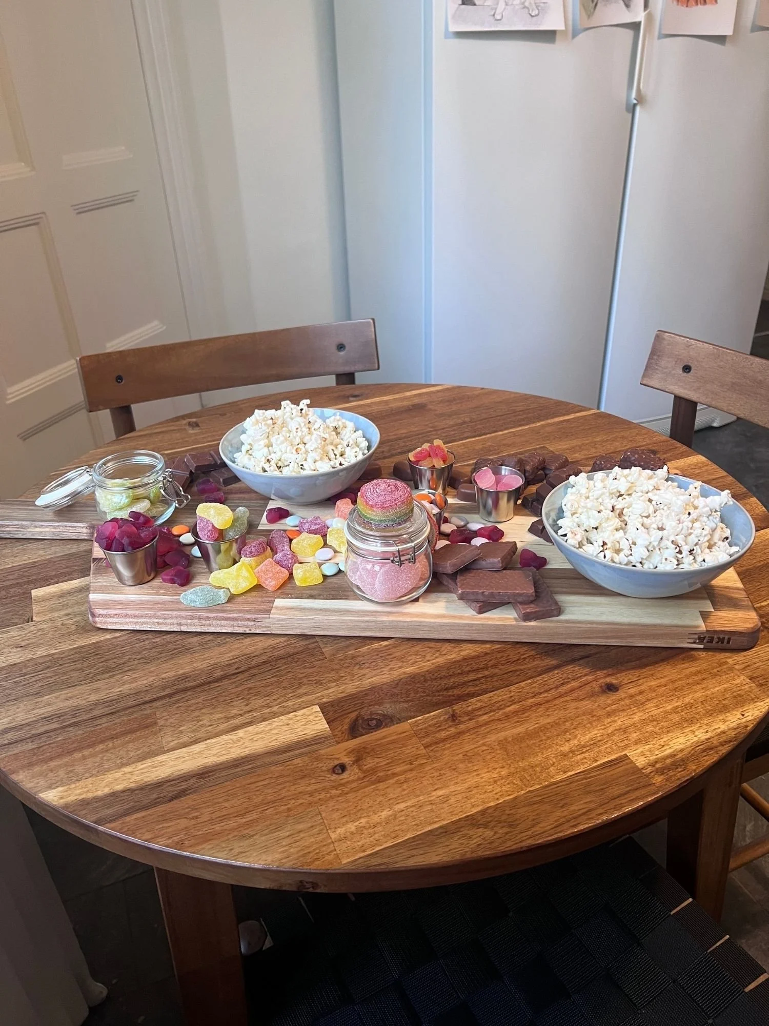 A wooden table set with a long snack board holding two bowls of popcorn, glass jars of gummies, small ramekins of candy, chocolate bars, and colorful sweets.