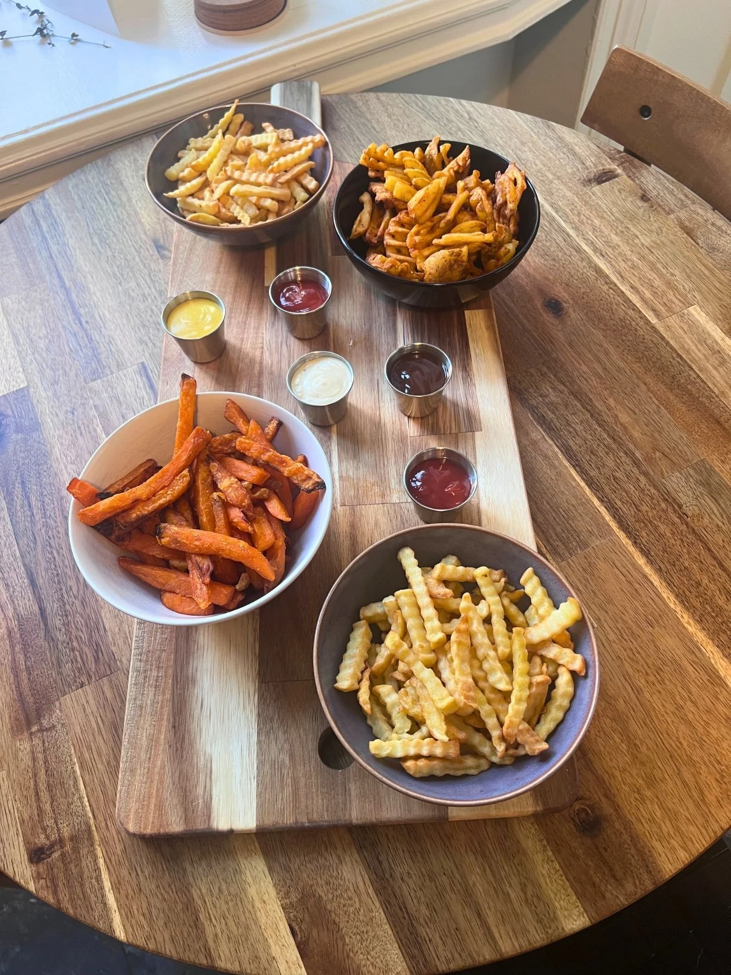 A long wooden board set on a round wooden table, holding four bowls of different style fries and sweet potato fries alongside six small metal ramekins of dipping sauces.