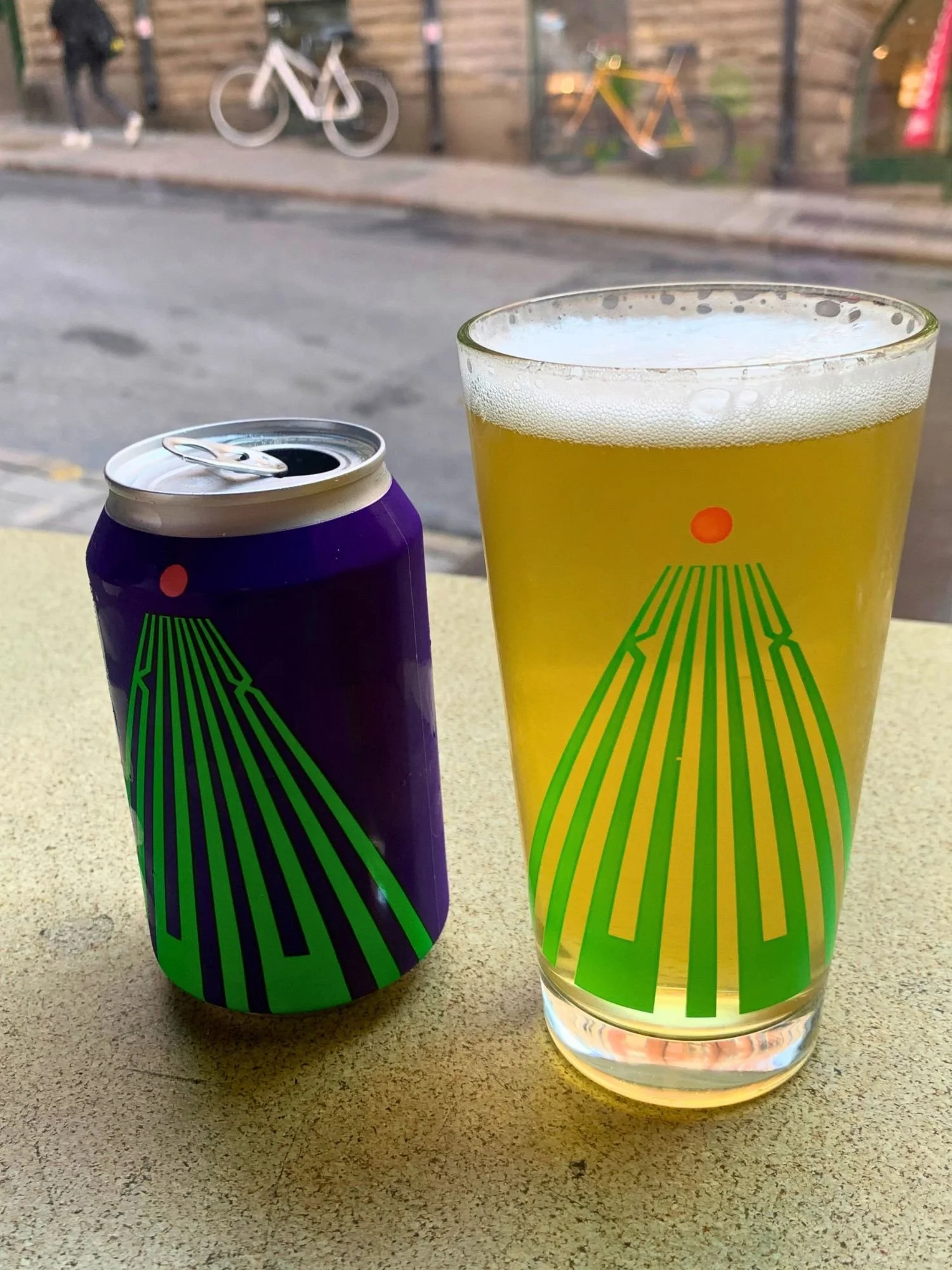 An open, deep blue beer can and a glass of hazy yellow beer on a bar. Both the glass and the can feature matching green graphic line designs topped with a small red dot.
