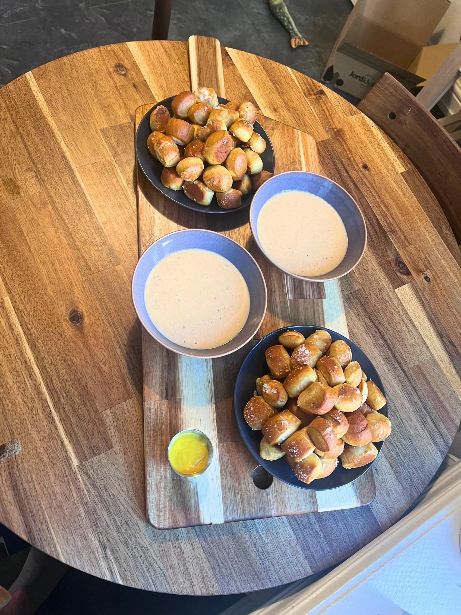 High-angle view of a wooden serving board set on a round wooden table, holding two plates of warm pretzel bites, two bowls of cheese dip, and a small metal ramekin of mustard sauce.