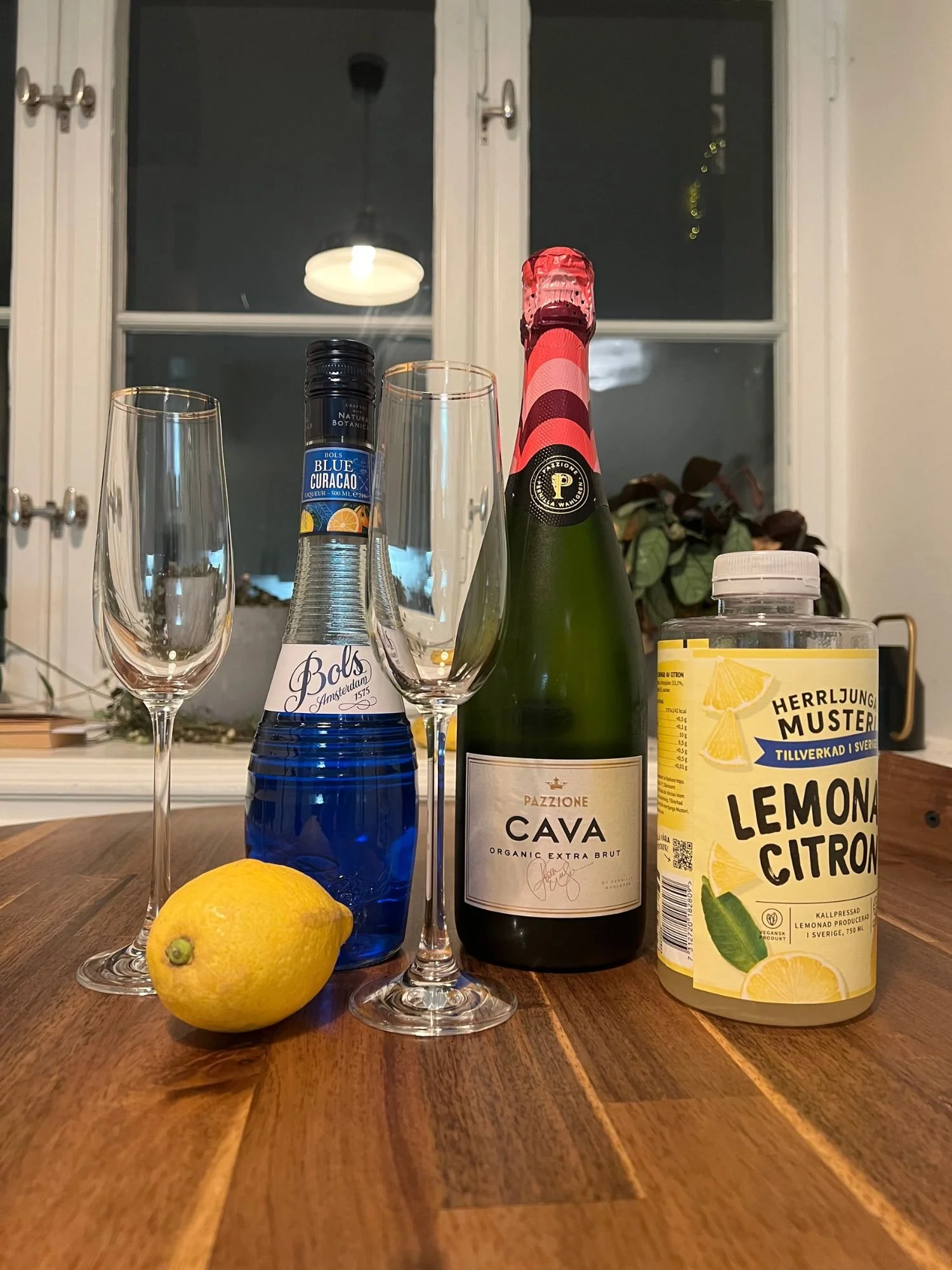 Ingredients for a blue cocktail on a wooden table, including blue curaçao, sparkling wine, lemonade, a lemon, and two champagne glasses, with a window and plants in the background.