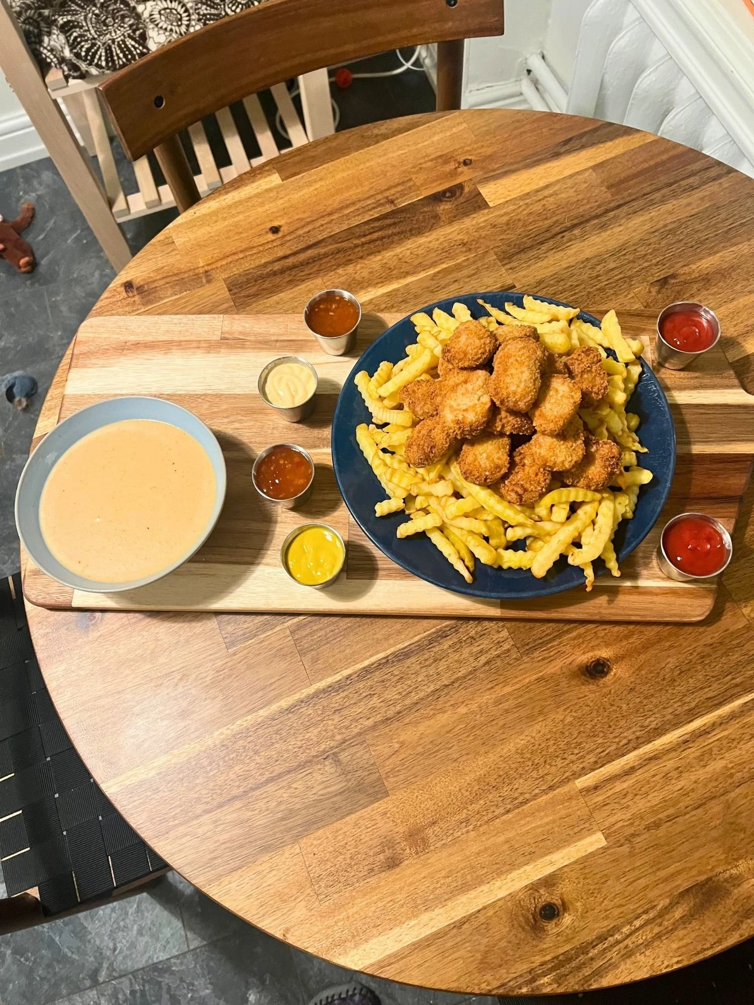 A large wooden serving board rests on a round wooden table, featuring a dark blue plate stacked high with golden crinkle-cut French fries and crispy fried nuggets.