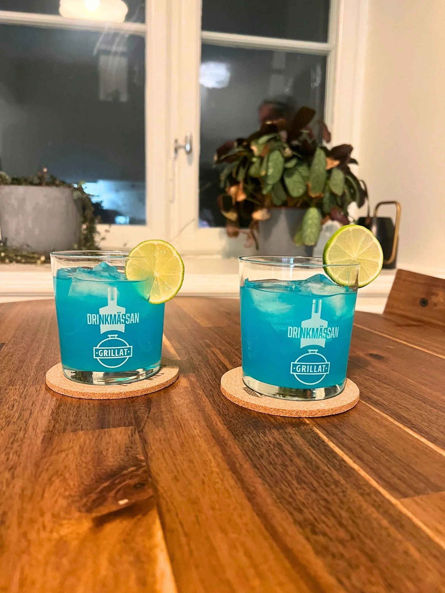 Two glasses of bright blue cocktails over ice, each garnished with a lime slice, placed on a wooden table with a window and houseplants in the background.