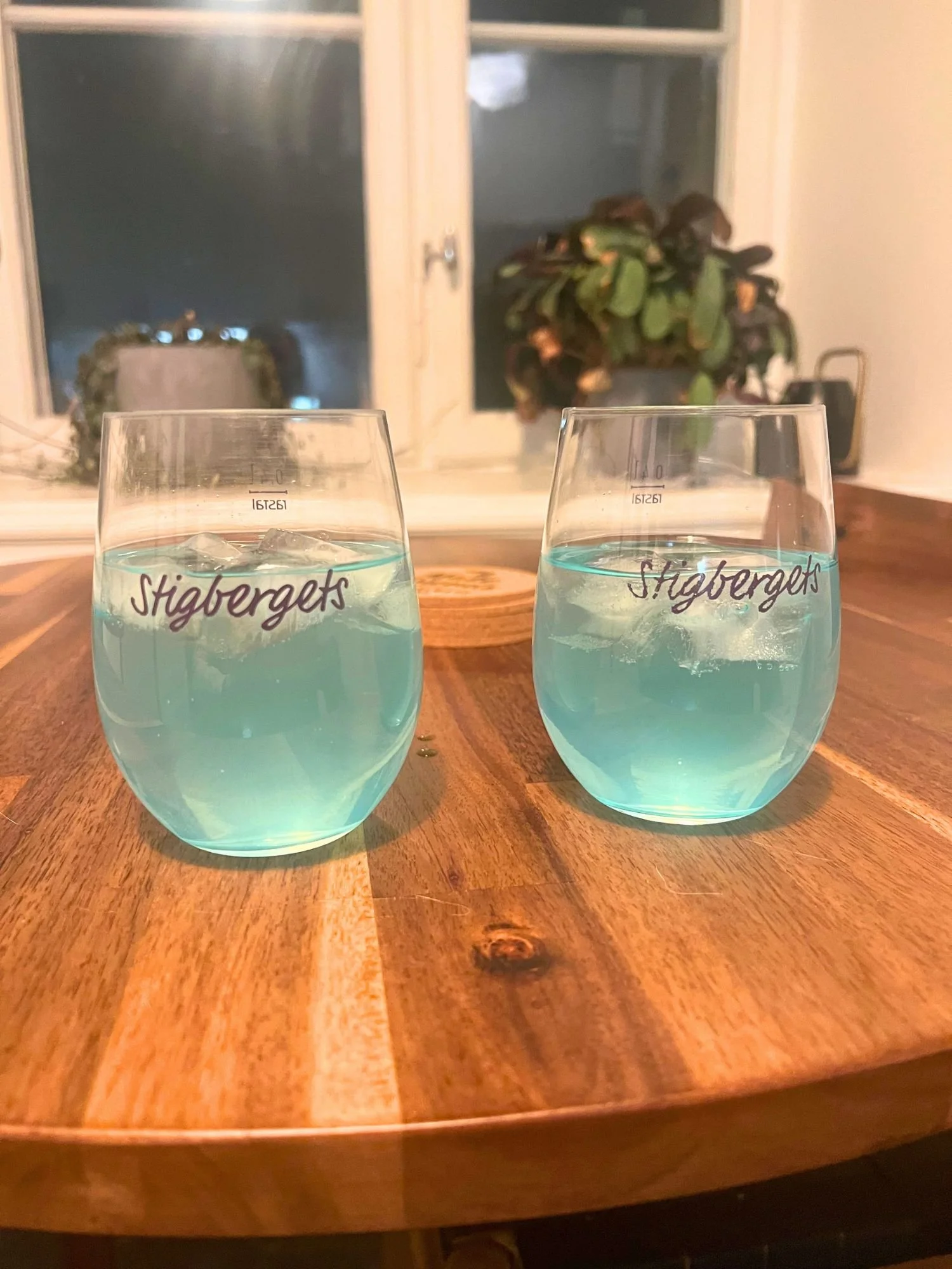 Two glasses of light blue cocktails with ice on a wooden table, with a window and houseplants in the background.