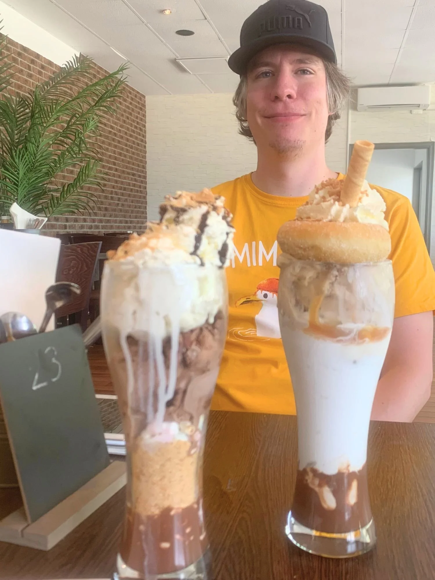 A smiling man in a yellow t-shirt and black cap sitting behind two giant, over-the-top milkshakes at a restaurant.