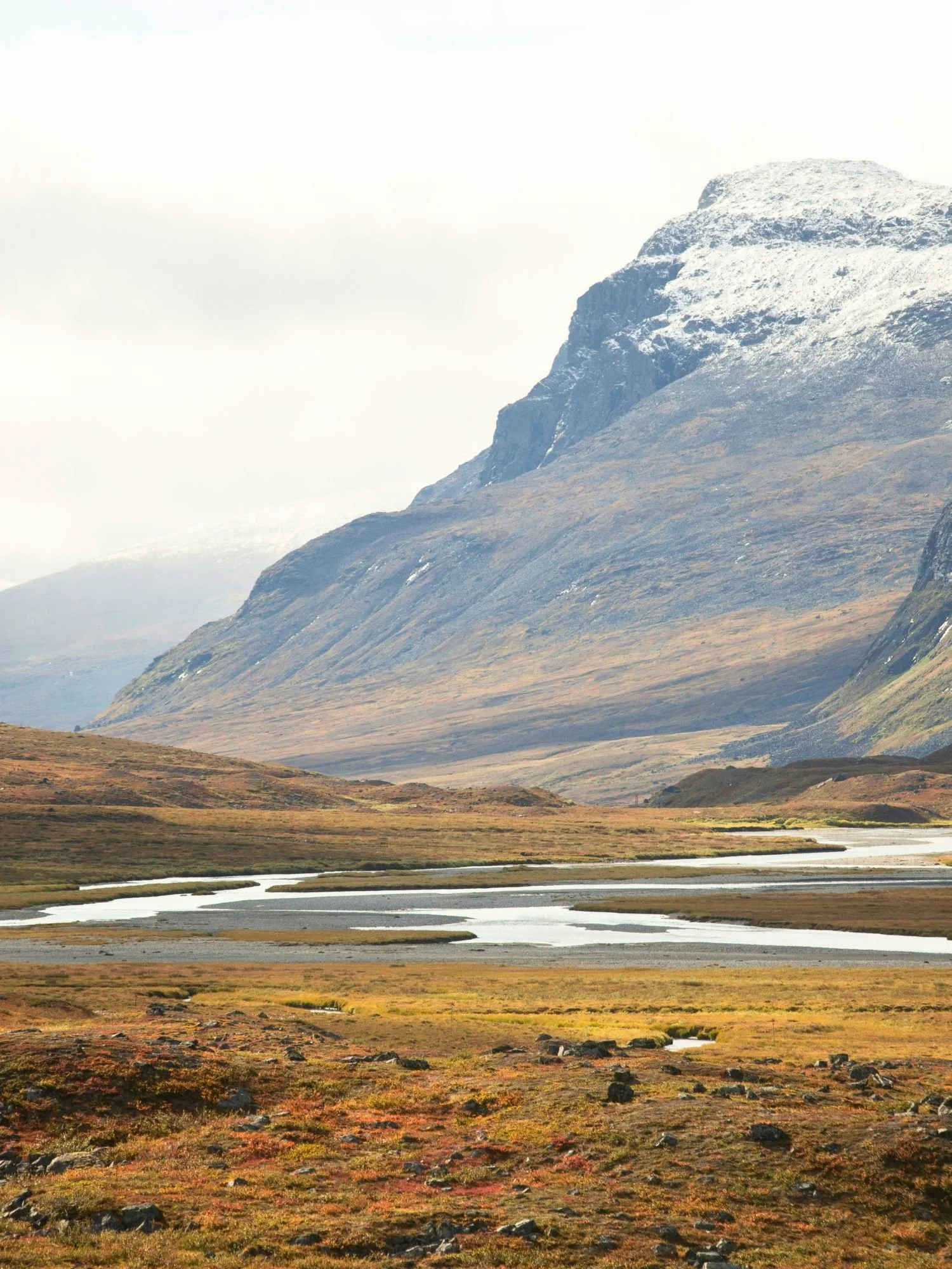 A vast mountain valley under a bright, overcast sky. In the foreground, the autumn tundra is a patchwork of gold, orange, and reddish-brown vegetation scattered with small dark rocks.