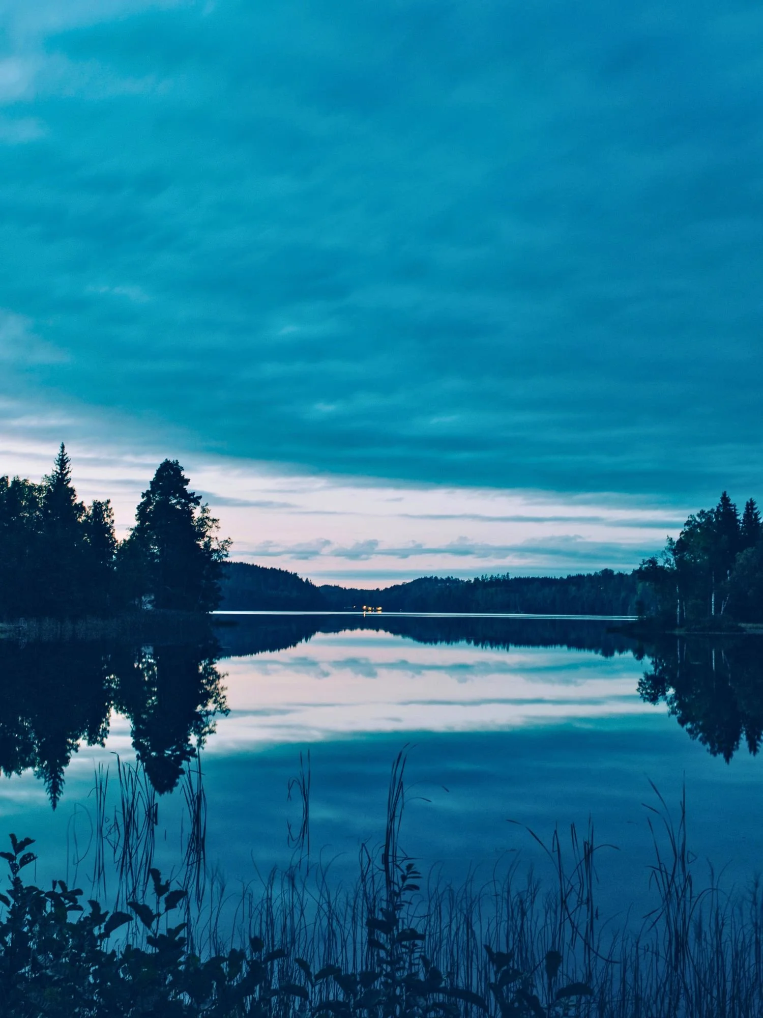A calm lake at twilight, in deep, moody shades of teal and blue. The glassy surface of the water mirrors the overcast sky and the dark silhouettes of pine forests lining the shore.
