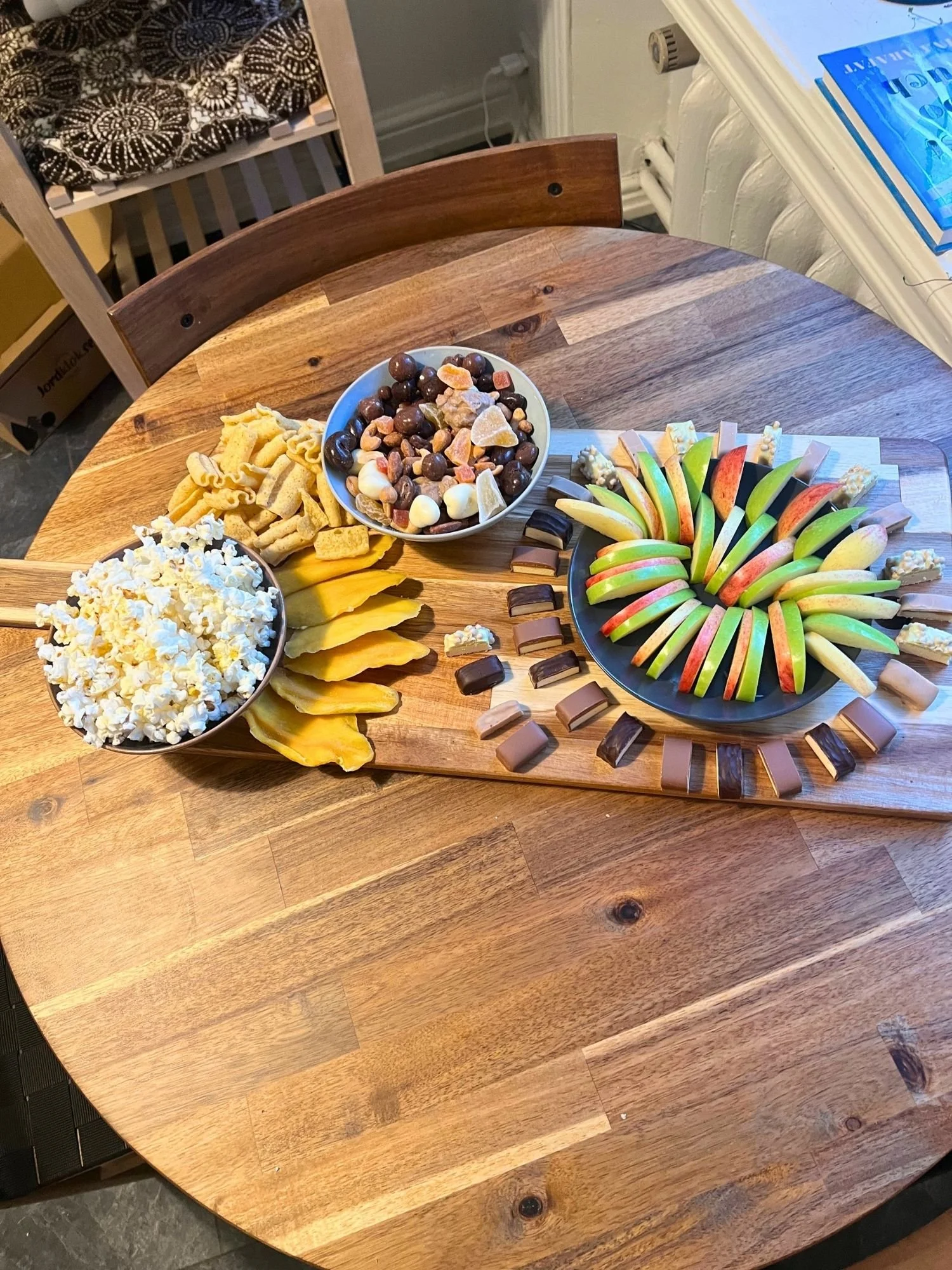 High-angle view of a wooden serving board set on a round wooden table, holding popcorn, trail mix, sliced dried mango, lentil chips, sliced apples, and protein bars.