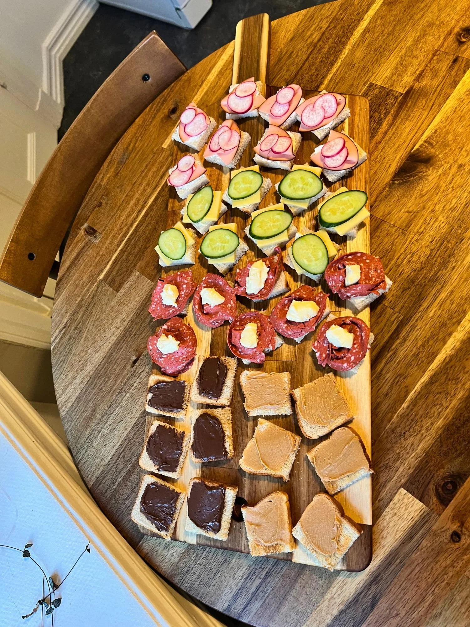 A long wooden board placed on a round wooden table, neatly arrayed with rows of small, square open-faced mini sandwiches.
