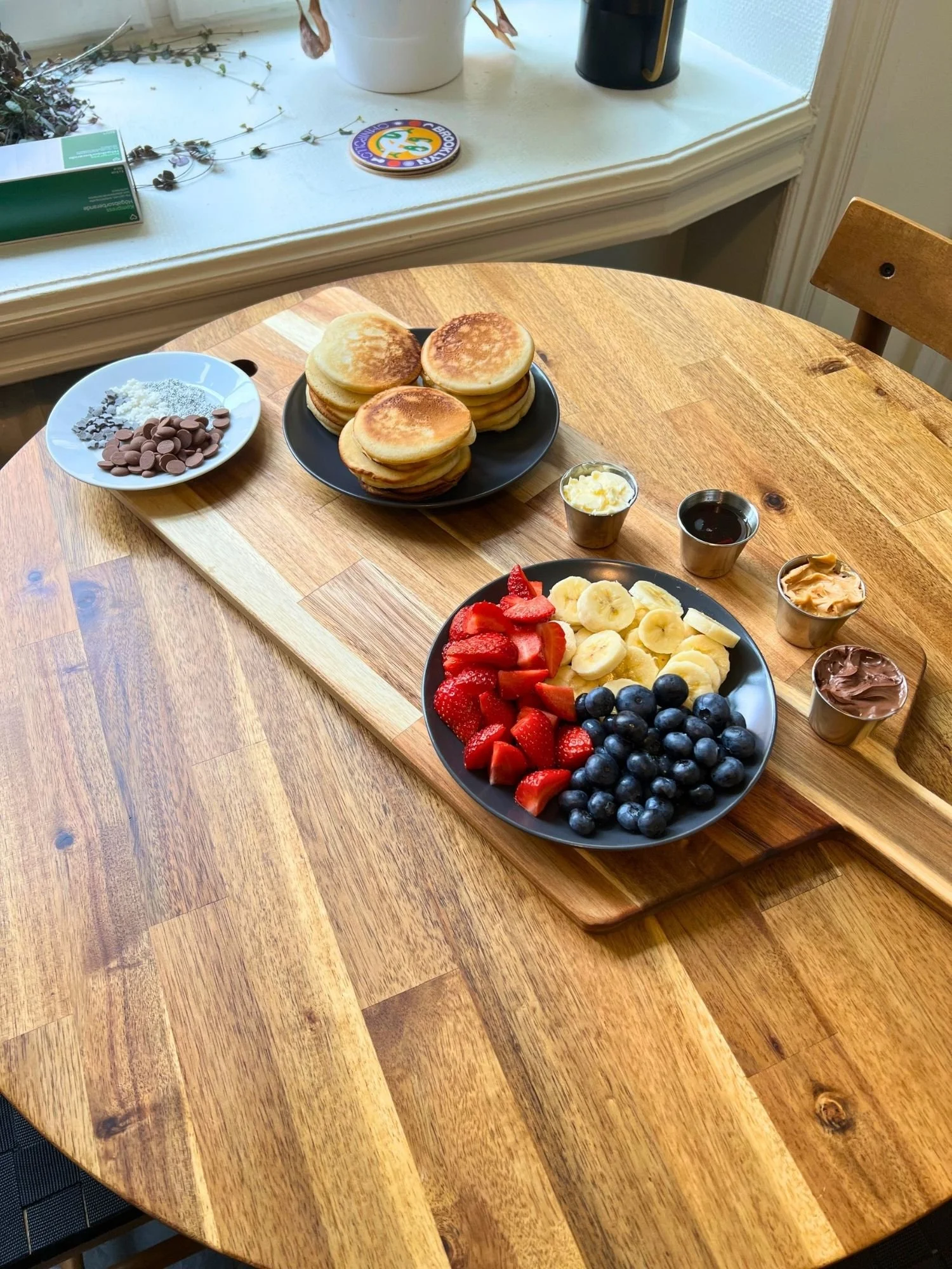 A long wooden serving board lies diagonally across a round wooden table, set with a mini pancake breakfast spread.