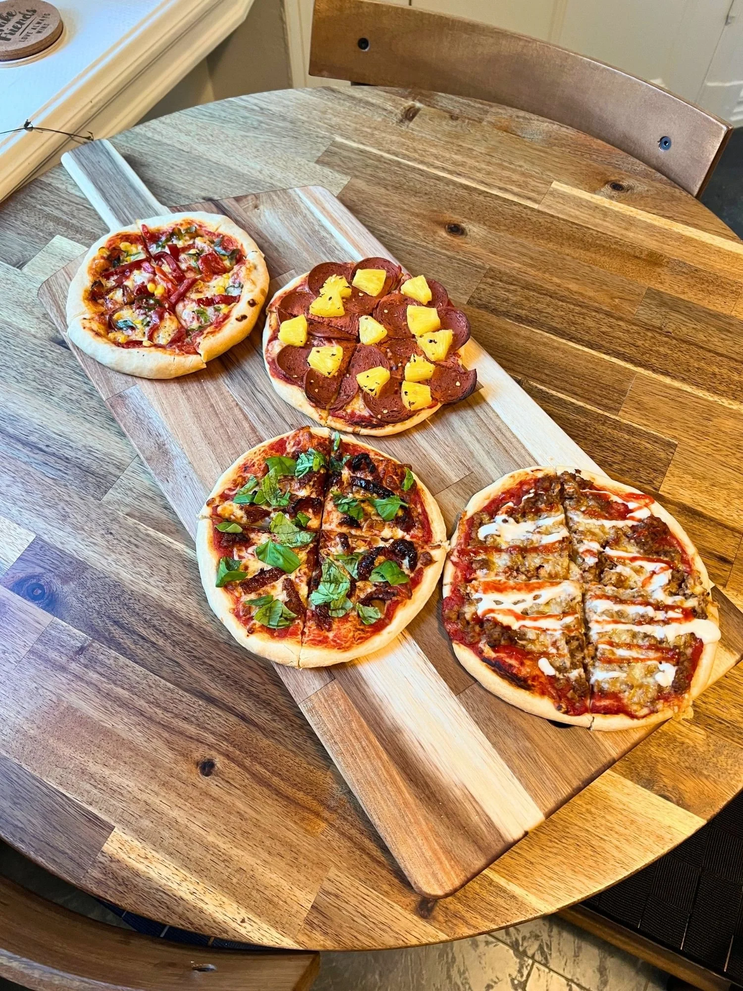 High-angle shot of a rectangular wooden serving board on a round wooden table, featuring four small personal-sized pizzas with different toppings.