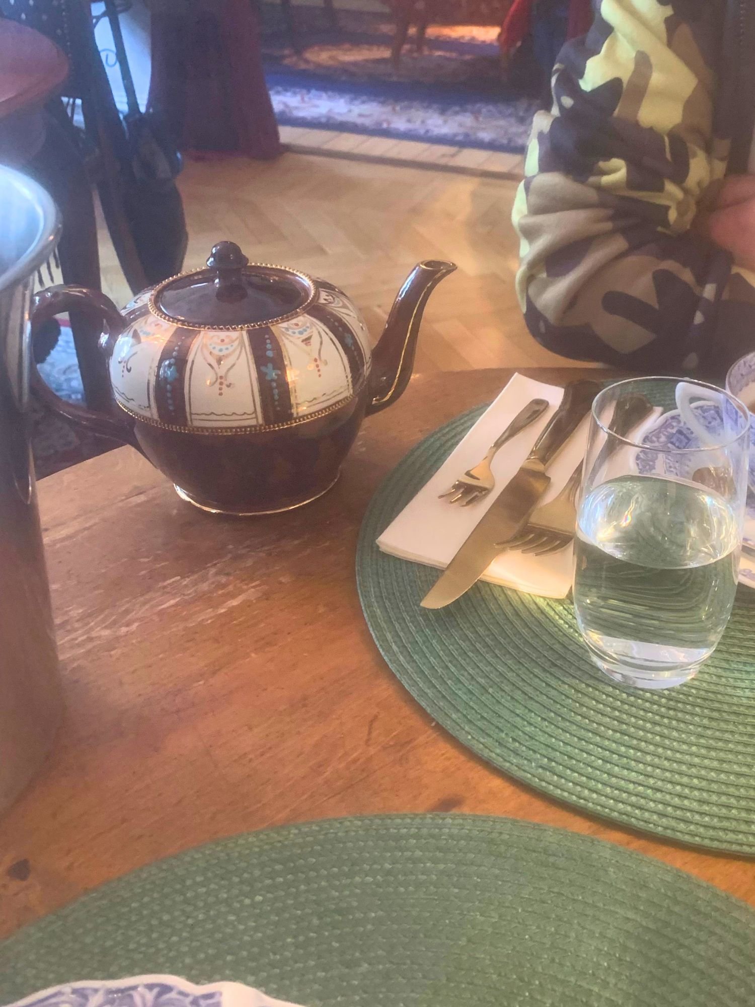 A tea setting on a wooden table at Alice och Kattens Tesalong in Lund. An ornate, dark brown ceramic teapot with a wide cream-colored band decorated with gold filigree and blue patterns.