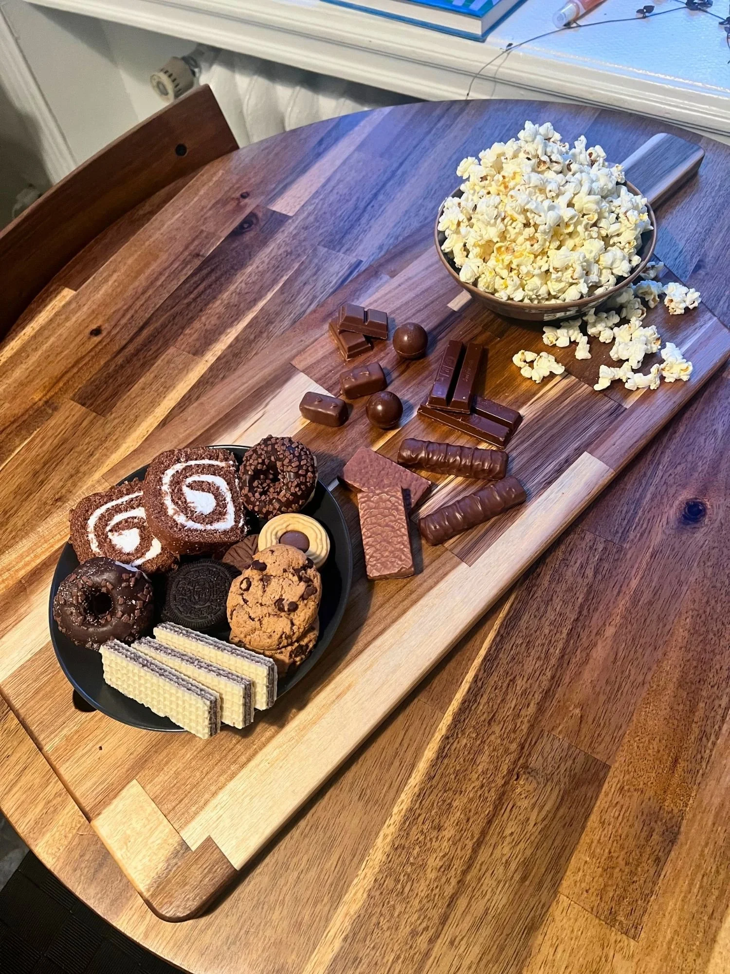 High-angle, diagonal view of a wooden serving board set on a round wooden table, holding a plate of cookies and mini donuts, assorted chocolates, and a bowl of popcorn.