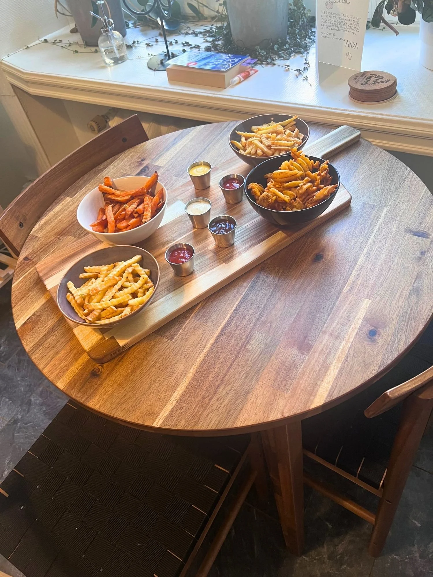 A wooden table topped with a long serving board containing four bowls of different styles of fries and sweet potato fries, along with five small metallic ramekins filled with various dipping sauces.