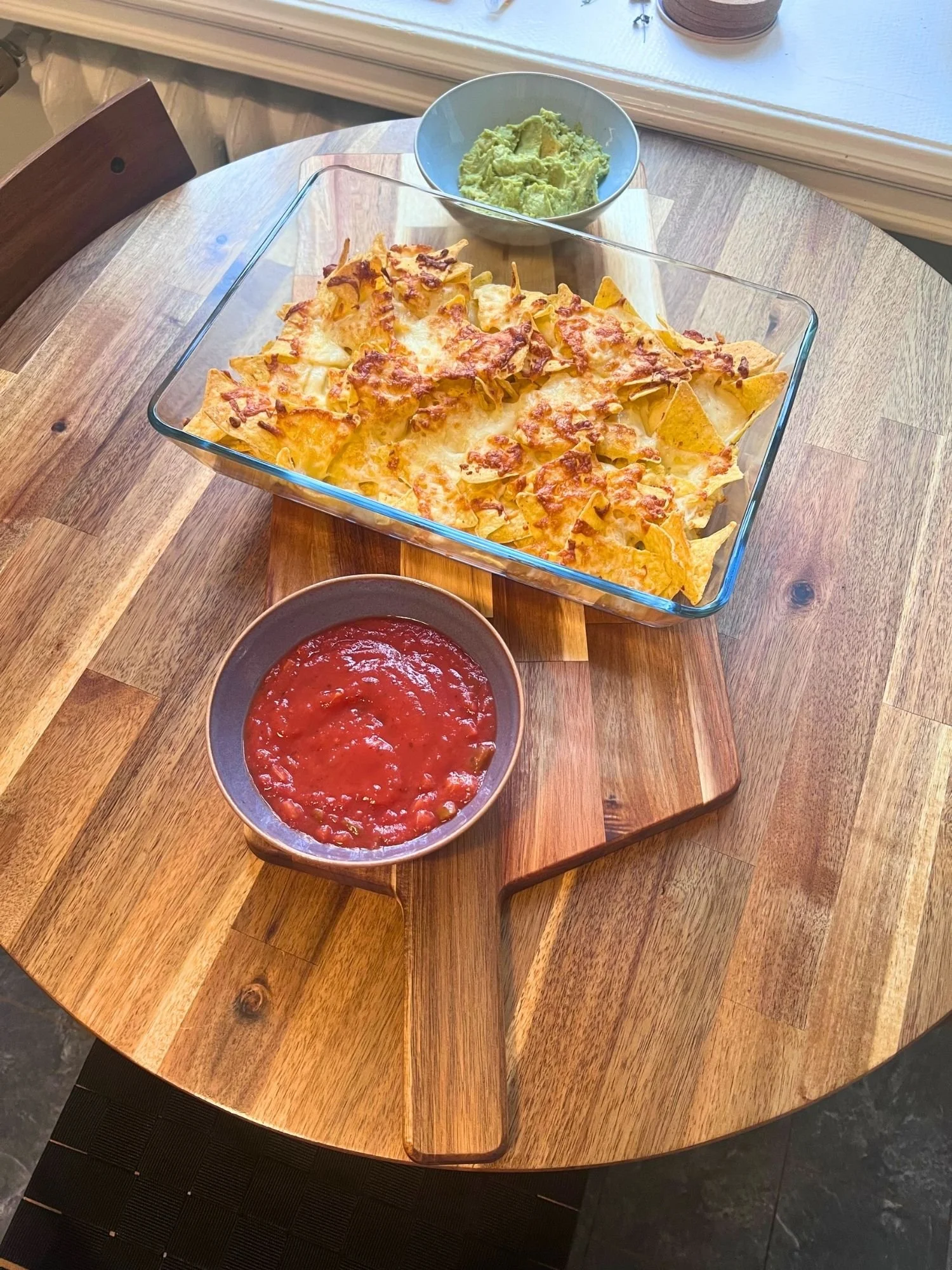 High-angle, length-wise shot of baked cheese nachos in a glass baking dish set on a wooden serving board alongside bowls of red salsa and green guacamole.