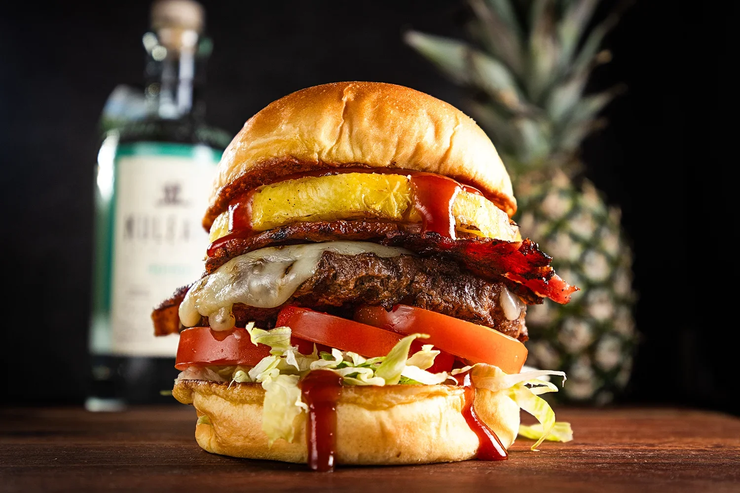 Hennessey's Kuleana Hawaiian Burger with grilled beef patty, melted cheese, bacon, grilled pineapple, lettuce, tomato, and sauce on a toasted bun, served on a wooden surface.
