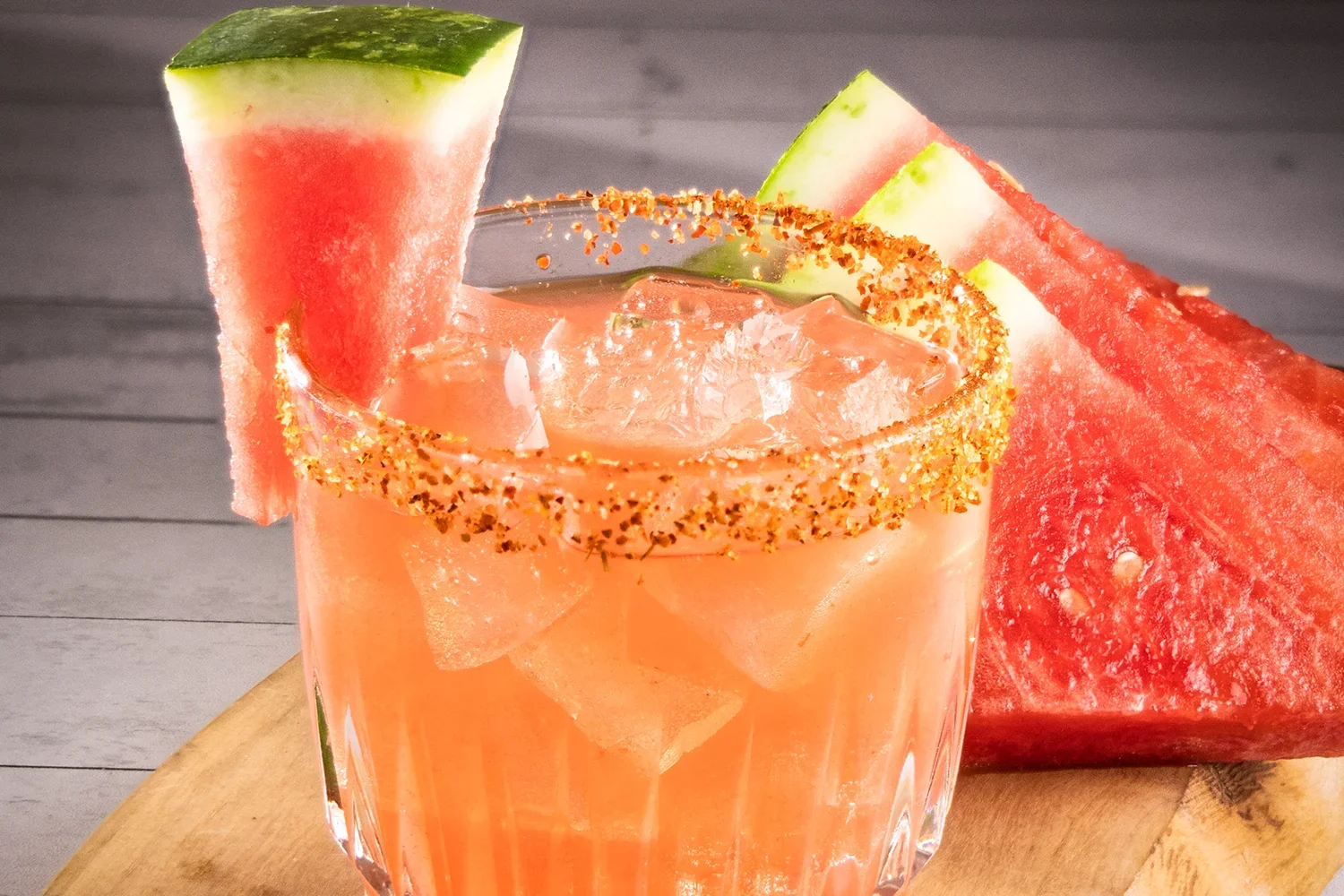 Hennessey’s Cocktail of the Month Lalomelon Margarita with ice, chili-salt rim, and watermelon garnish, served beside fresh watermelon slices on a wooden board.
