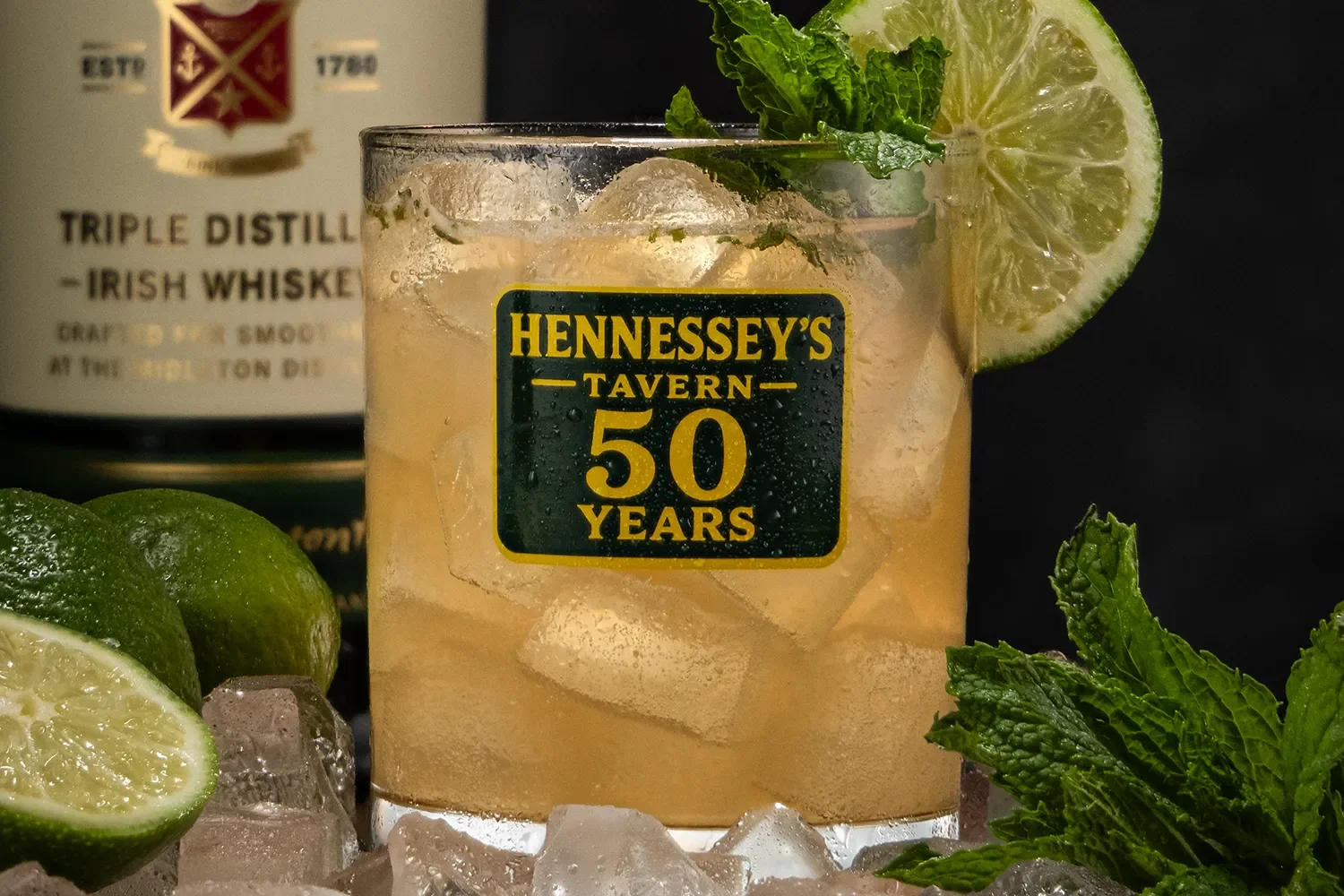 Close-up of the Half Century Highball over ice in a Hennessey’s Tavern 50 Years glass, garnished with lime and mint beside a Jameson bottle.