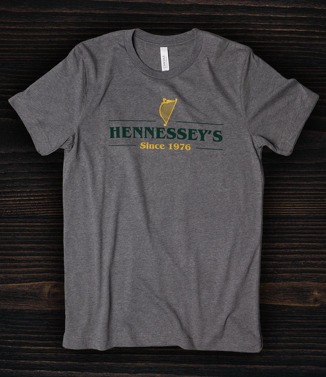 Gray Hennessey’s Tavern T-shirt featuring a green and gold logo with a harp graphic and “Since 1976” text, displayed against a dark wood background.