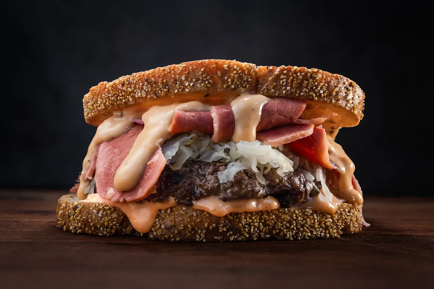 Hennessey’s Tavern O’Reuben Burger with 1000 island, corned beef, sauerkraut, burger patty, and beer cheese on seeded rye bread, close-up against a dark background.