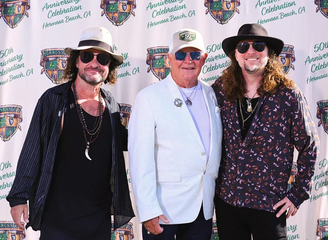 Three smiling men wearing hats and sunglasses pose in front of Hennessey’s Tavern’s 50th Anniversary backdrop in Hermosa Beach.