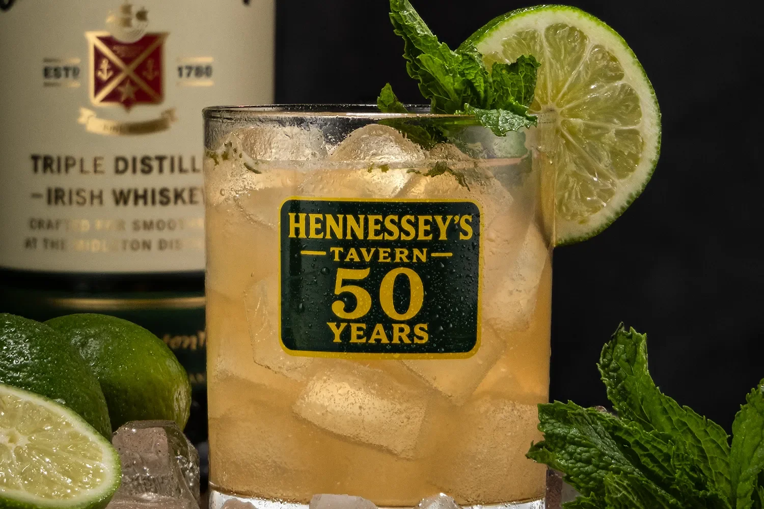 Half Century Highball garnished with a lime slice and mint, labeled "Hennessey's Tavern 50 Years," with limes, mint, and a whiskey bottle in the background.