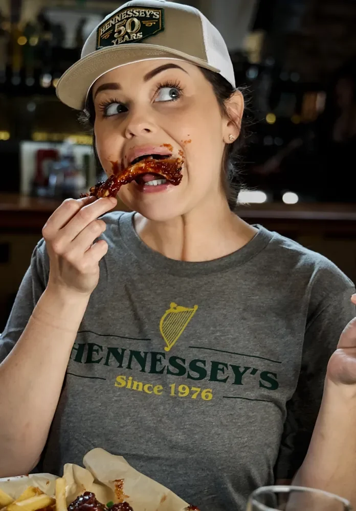 Woman in a Hennessey’s Tavern shirt and hat eats saucy ribs at a table with fries.