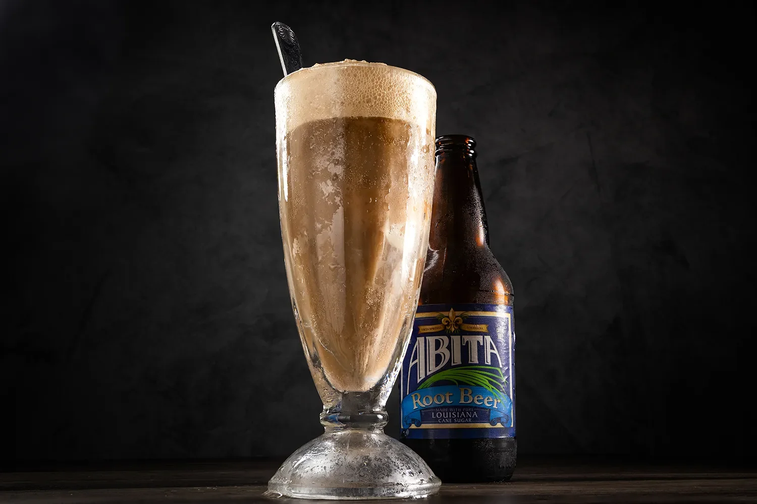 Root beer float in a glass with foam and a spoon, next to a bottle of Abita Root Beer on a dark background.