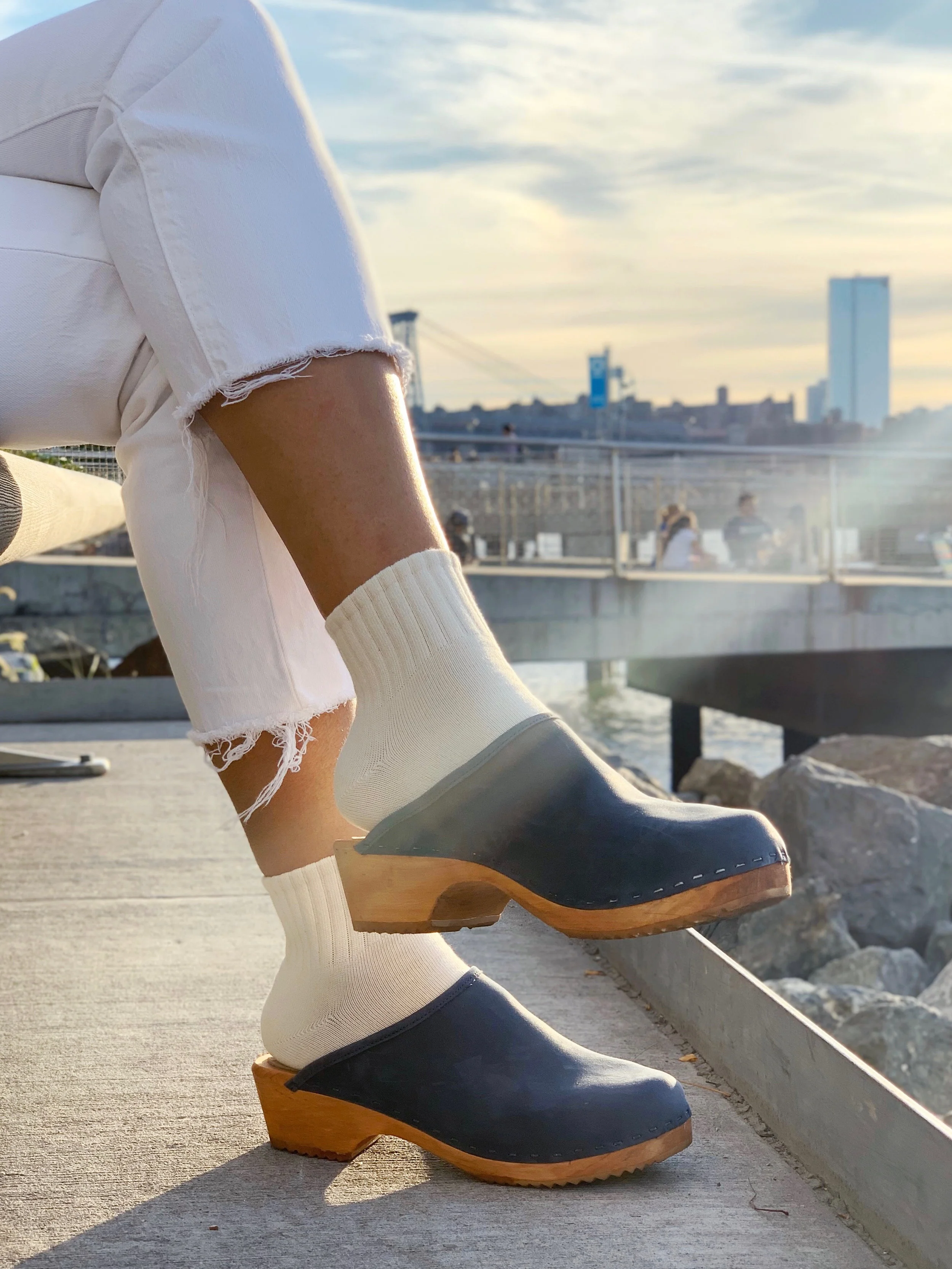 White jeans, cream socks, and dark blue Tessa clogs photographed in front of city skyline/waters edge