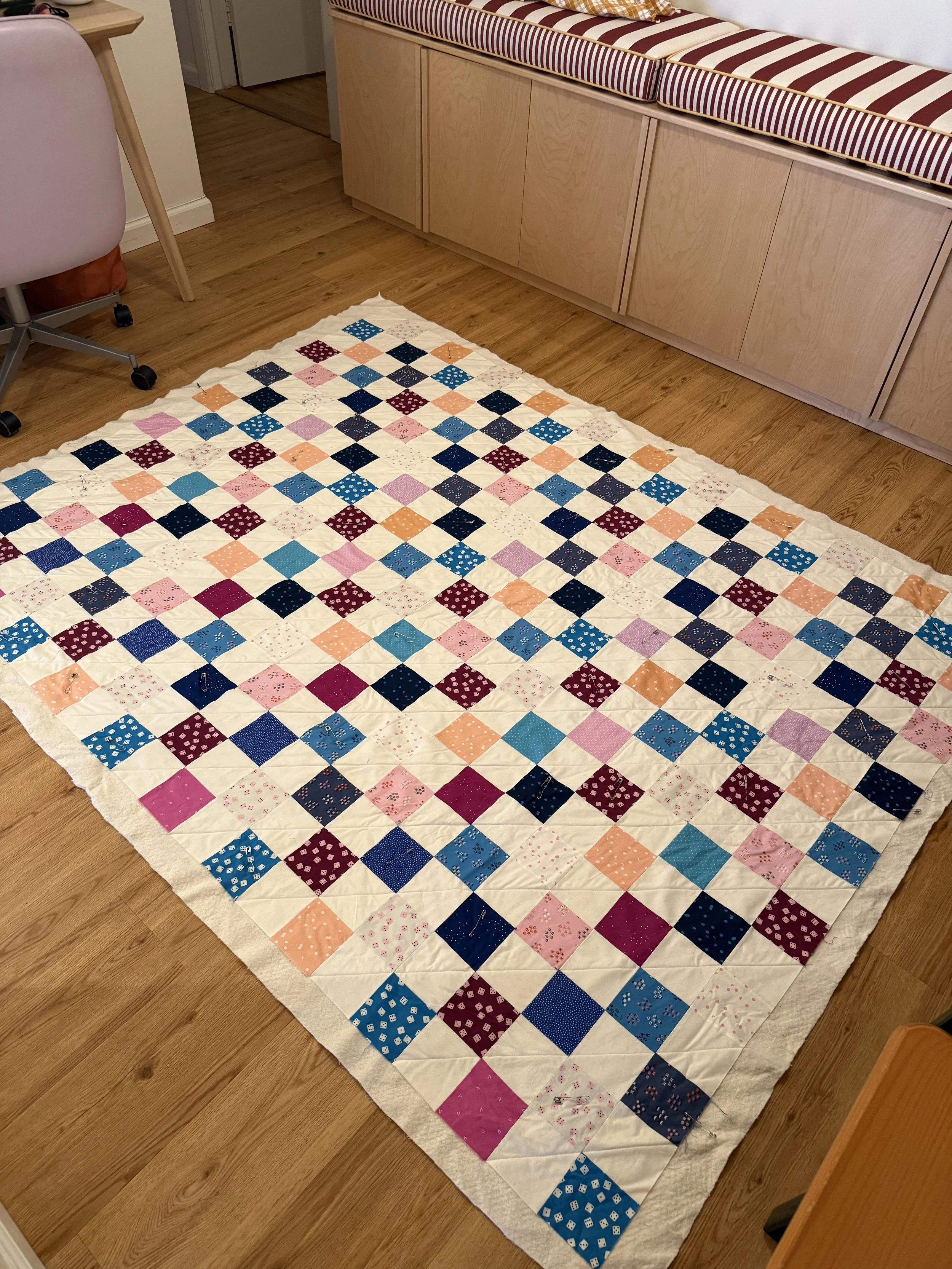 A patchwork quilt with mixed fabrics in blue, pink, peach, and maroon shades laid out on an apartment floor with the inner batting visible