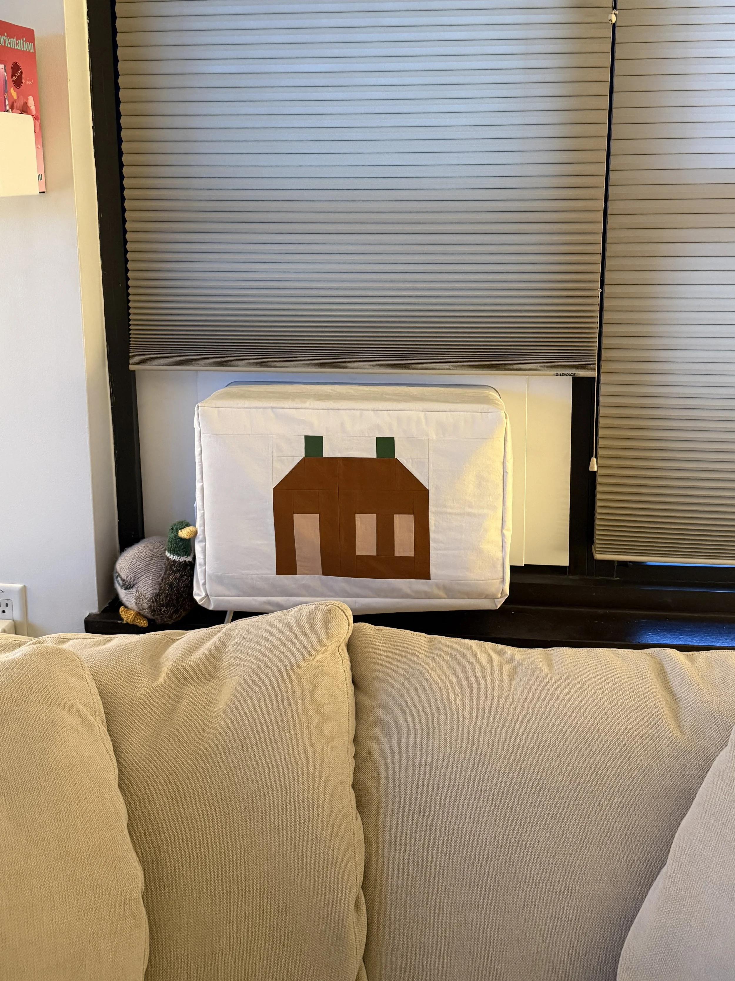A New York City apartment window with a window air conditioner unit. The unit is covered in a DIY cover that features a brown home with green chimneys
