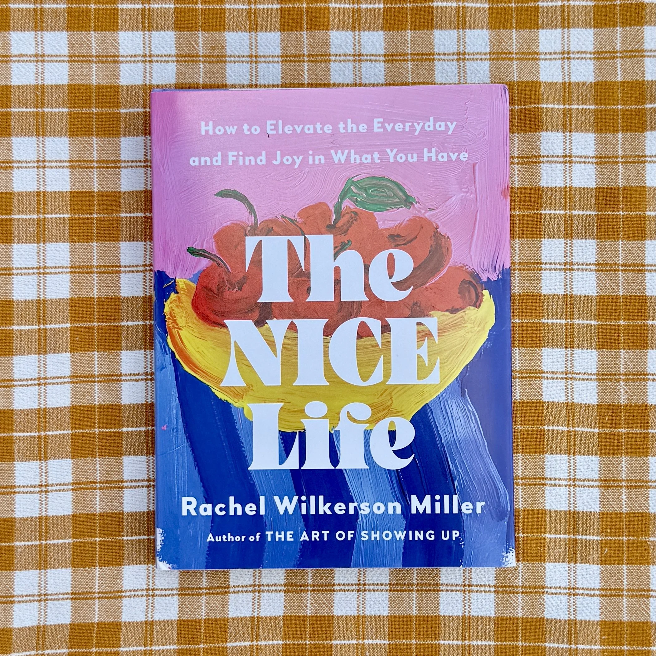 The Nice Life: How to Elevate the Everyday and Find Joy in What You Have by Rachel Wilkerson Miller. The book cover shows a hand-painted still life with a pink upper, a bowl of cherries in a yellow bowl on top of a blue striped tablecloth.