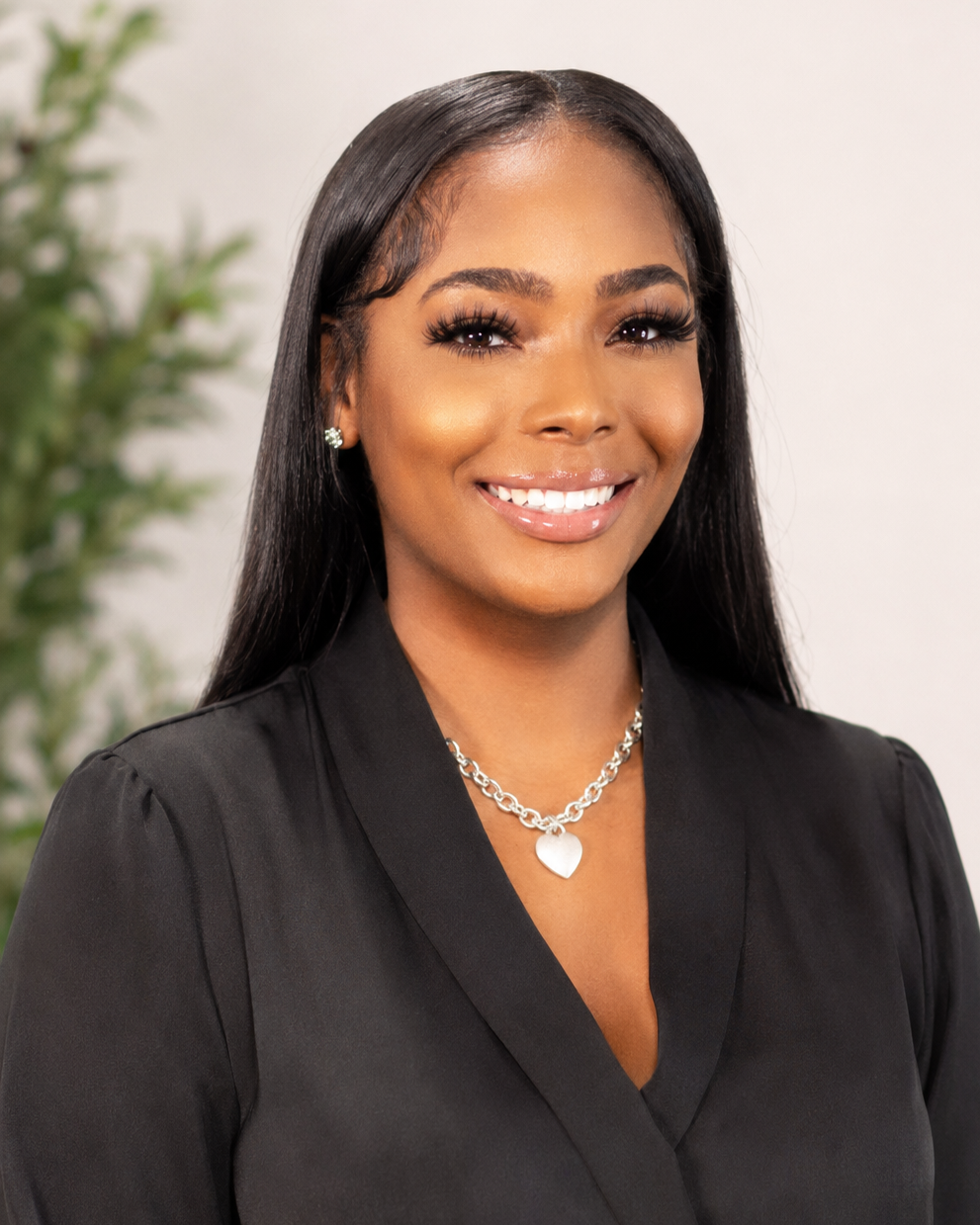 Portrait of a smiling woman with long black hair wearing a black blazer, silver heart necklace, and earrings, standing against a light background with a blurred green plant in the background.