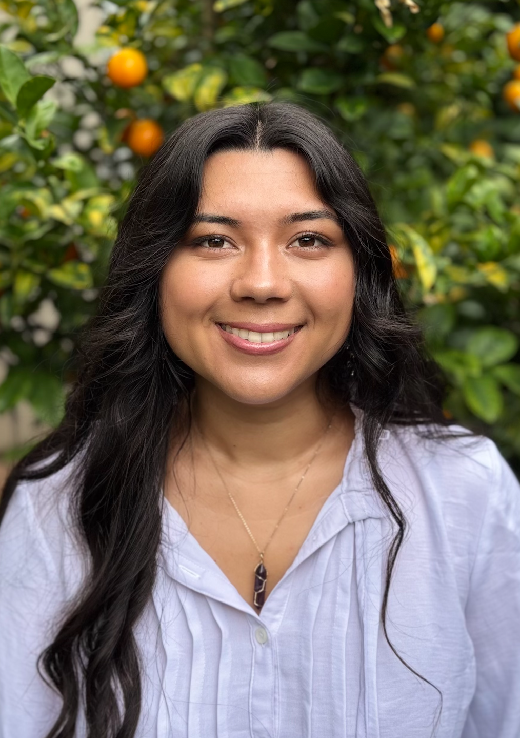 A woman smiling outdoors with greenery and orange fruit in the background.