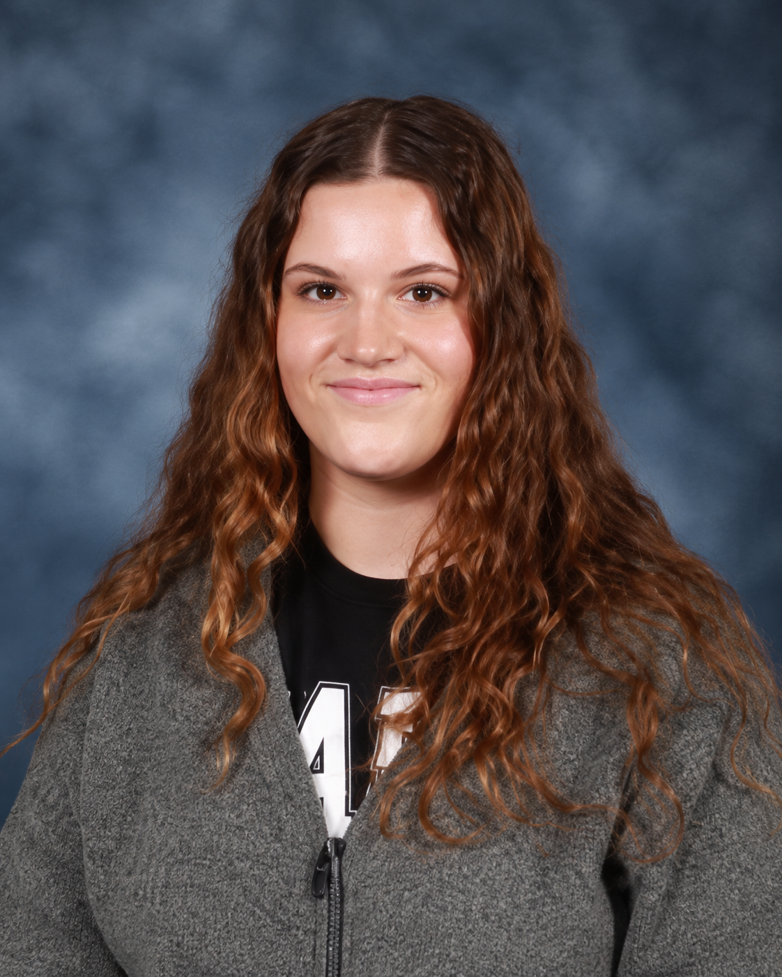Young woman with long, wavy brown hair, smiling, wearing a gray zip-up jacket and black shirt with white lettering, against a blue-gray background.