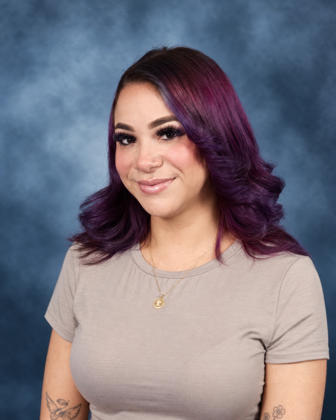 A young woman with purple ombre hair, wearing a beige t-shirt and a gold necklace, smiling against a blue background.