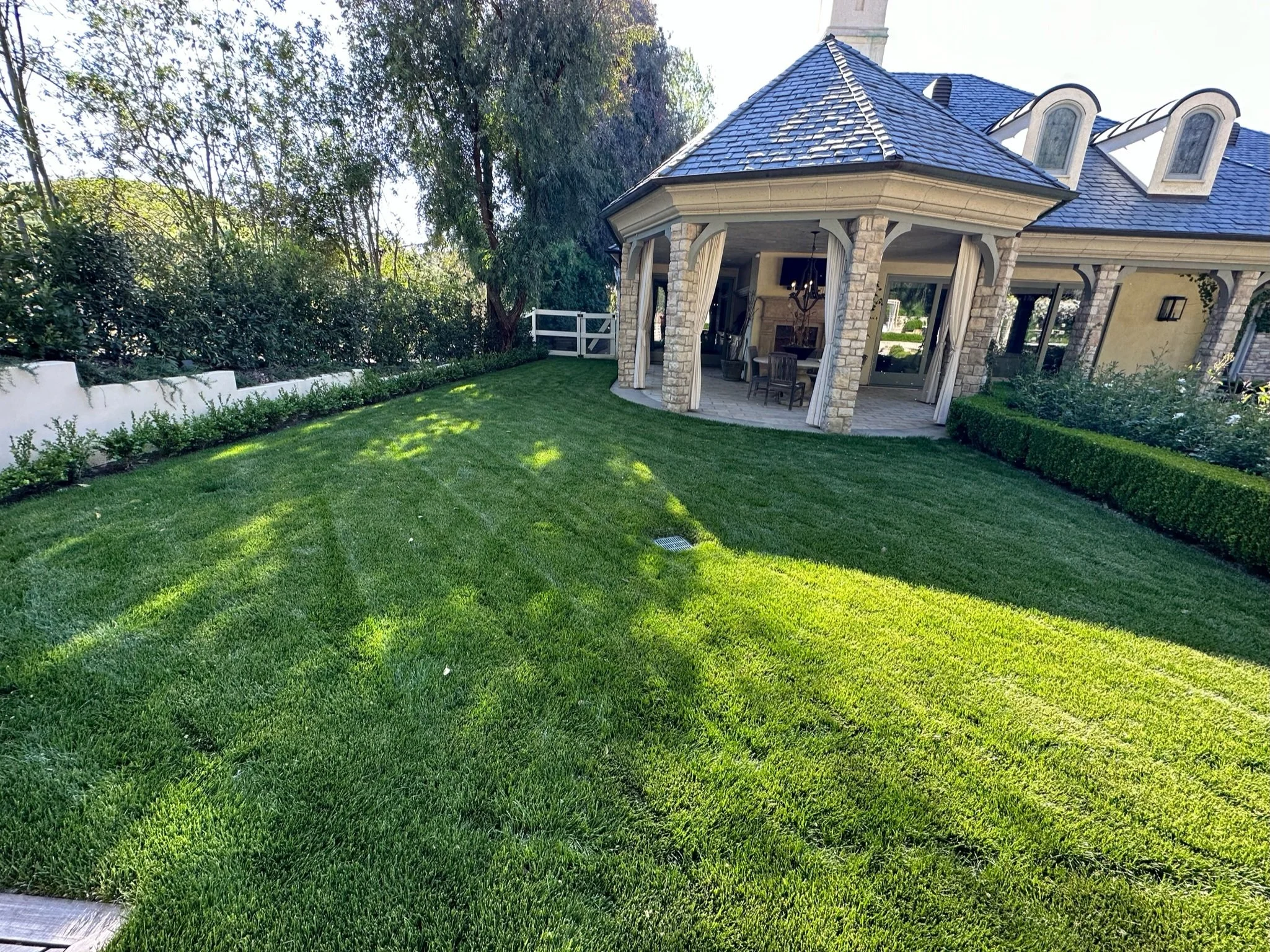 A well-maintained backyard with lush green grass, a small patio area with chairs and a chandelier, and surrounding bushes and trees. The house has a stone exterior and a blue shingled roof.