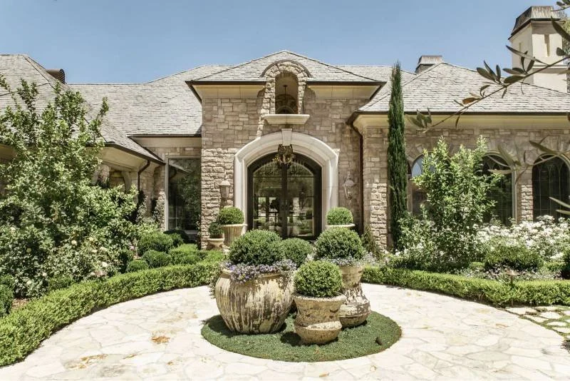 Front entrance of a large stone house with a well-maintained garden and potted plants.