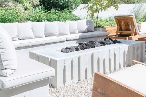 Outdoor seating area with white cushions, a concrete fire pit with rocks, and wooden furniture under a partially shaded area.