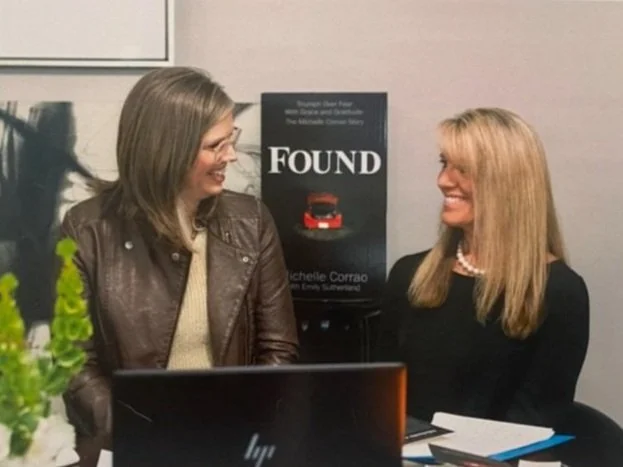 Two women sitting at a table, smiling and talking, with a poster titled 'FOUND' and a laptop in front of them.